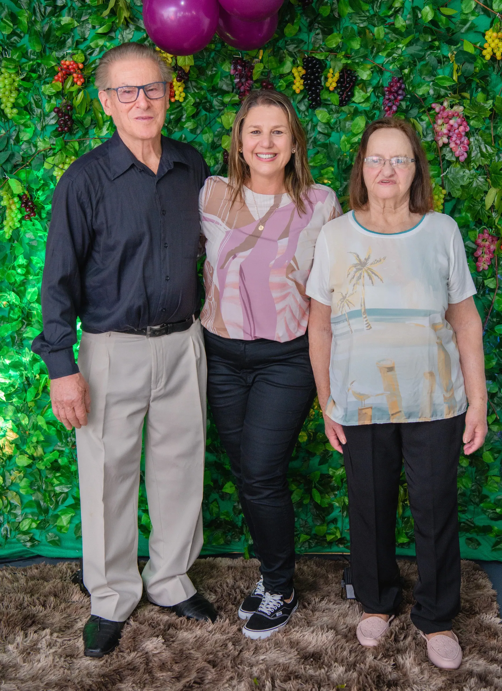Três pessoas, um homem e duas mulheres, em pé e posando para uma foto na frente de uma parede coberta de videiras falsas com cachos de uvas roxas e vermelhas.