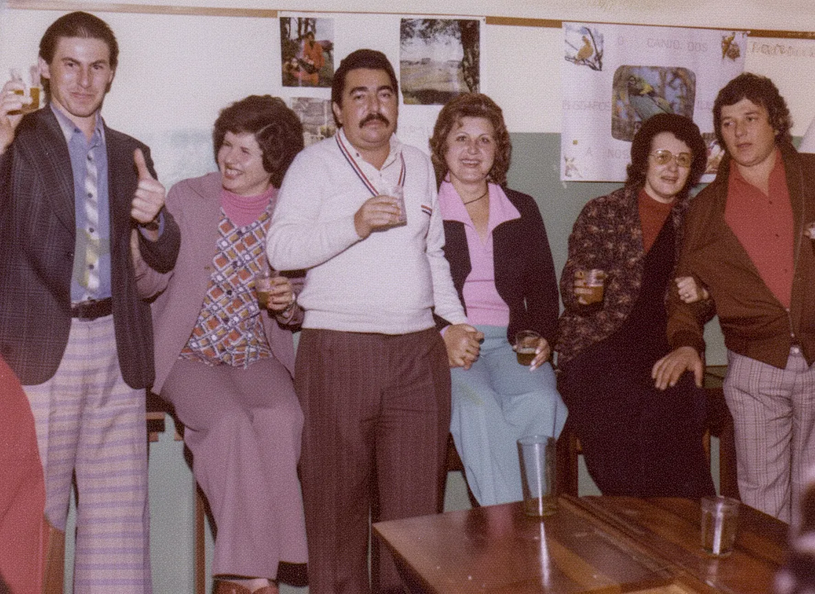 Grupo de seis adultos com bebidas em uma sala de aula de estilo antigo, com paredes brancas, pôsteres infantis e uma mesa de madeira escura com copos e um pires.