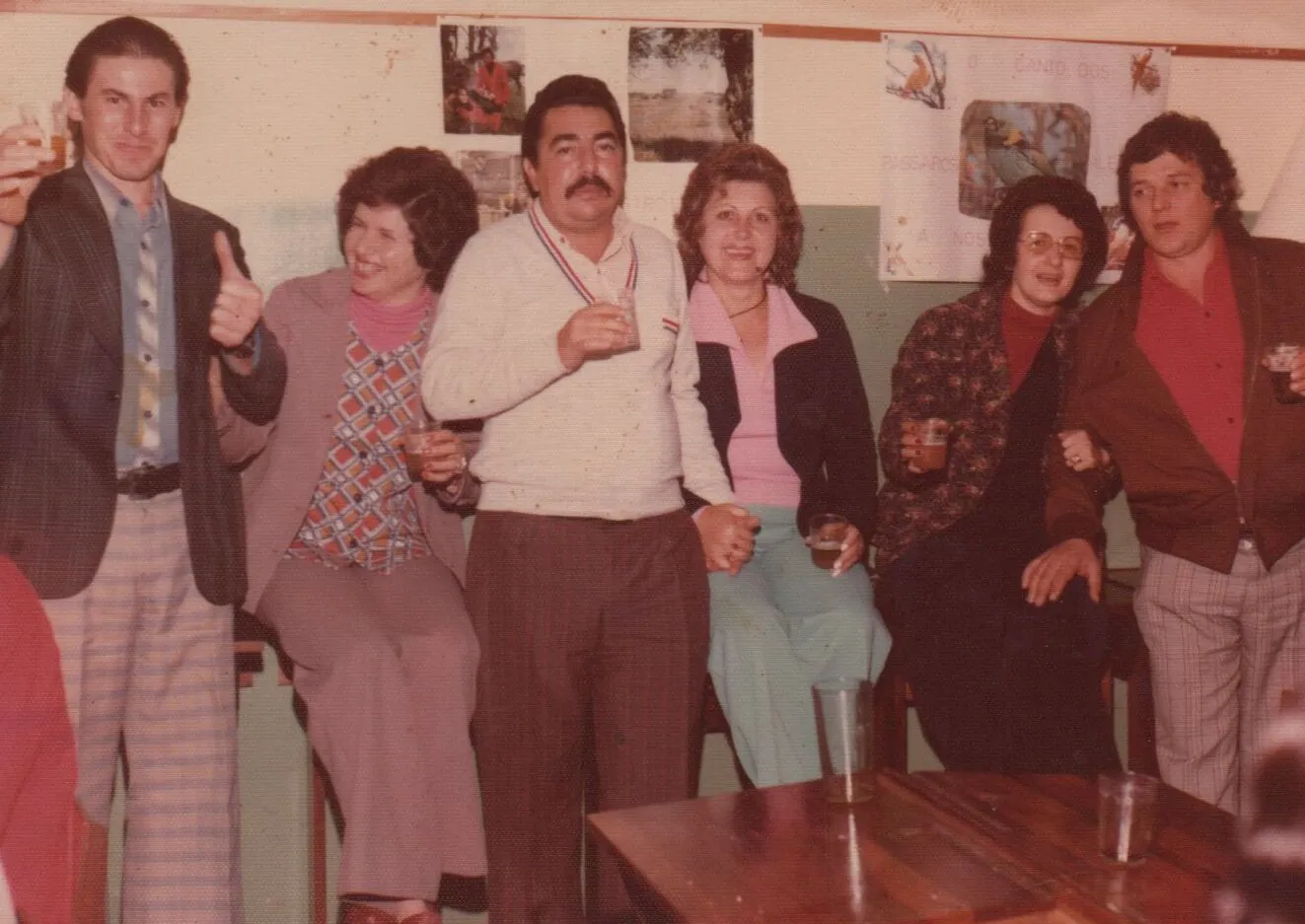 Grupo de seis adultos com bebidas em uma sala de aula de estilo antigo, com paredes brancas, pôsteres infantis e uma mesa de madeira escura com copos e um pires. (Original)