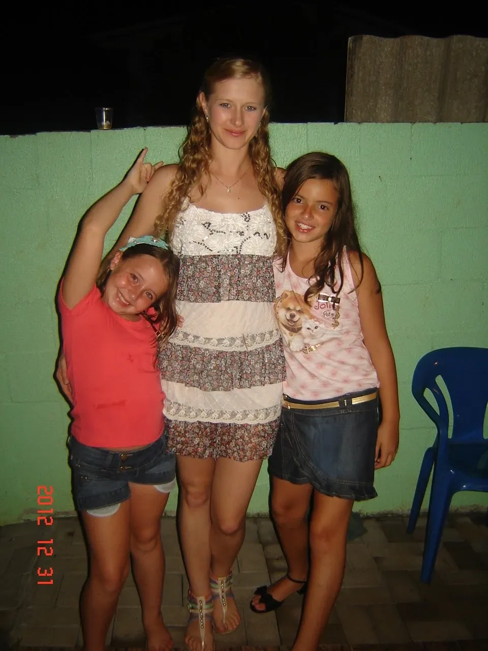 Uma mulher loira sorrindo com um vestido floral sem alças abraça duas meninas sorrindo; a menina à esquerda em uma camiseta vermelha e shorts jeans e a menina à direita em uma camiseta rosa e saia jeans.