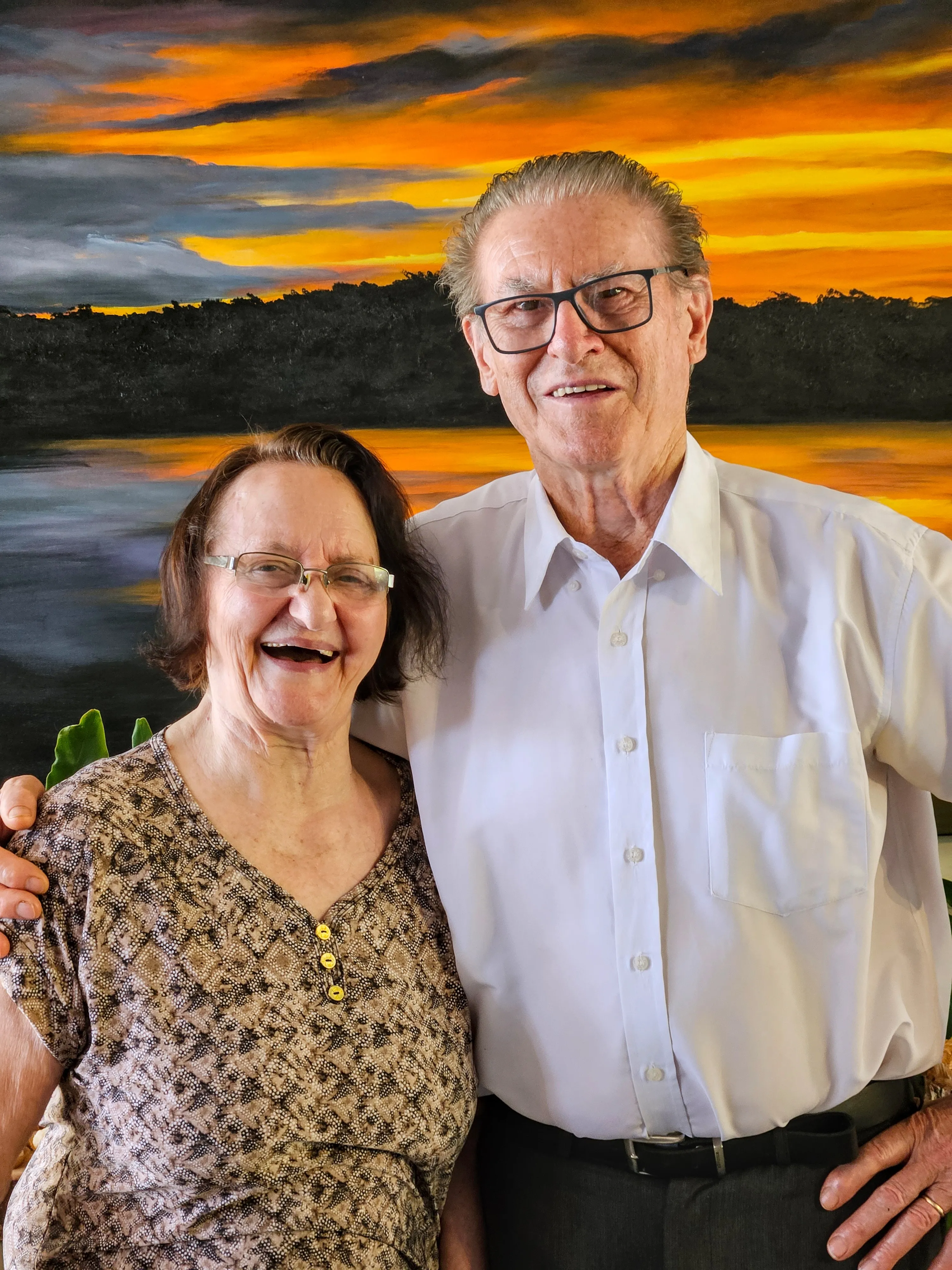 Um casal idoso sorri para a câmera, com o braço do homem em volta da mulher, contra um pano de fundo de pintura de paisagem ao pôr do sol.