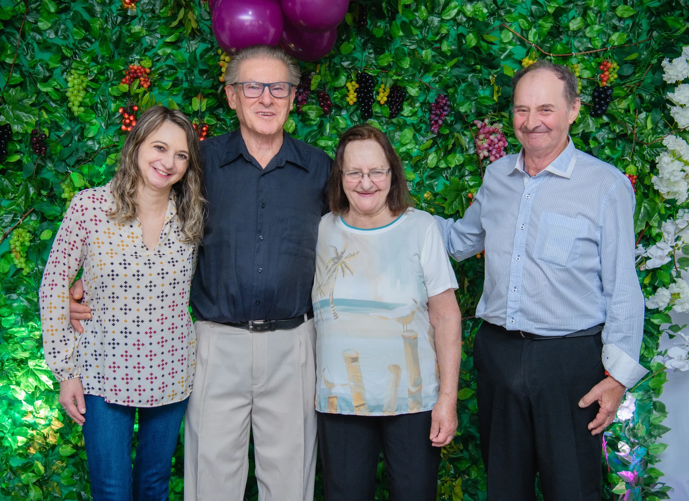 Quatro adultos - duas mulheres e dois homens - posam juntos em frente a uma parede de folhagem com uvas e balões roxos.