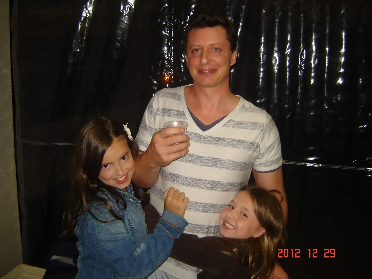 Um homem sorridente com aparelho nos dentes, vestindo uma camiseta listrada, segura um copo enquanto abraça duas meninas sorridentes, uma à sua esquerda com uma jaqueta jeans e outra à sua direita, ambas com cabelo castanho.