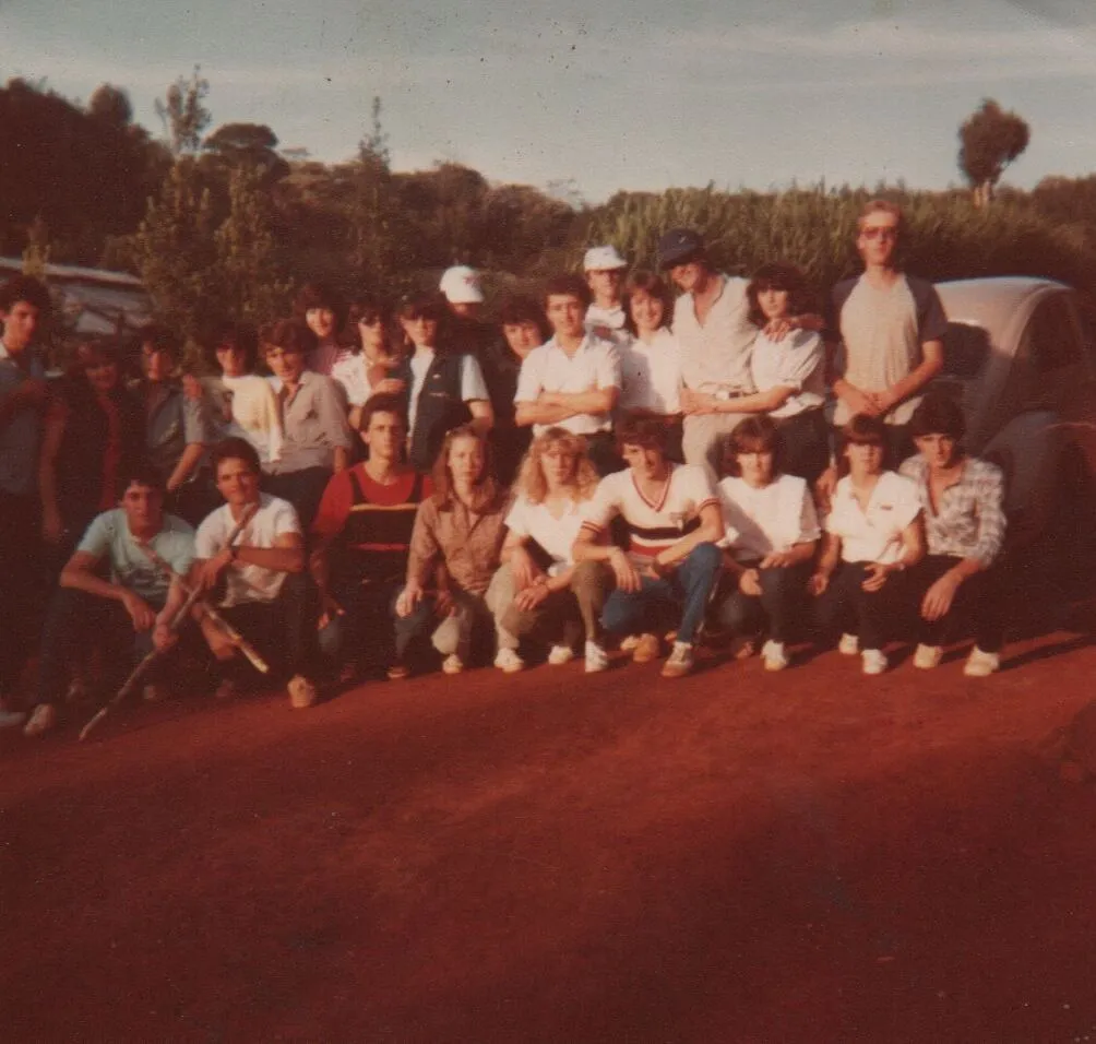 Um grupo de pessoas posando para uma foto ao ar livre, algumas sentadas e outras em pé, com árvores e um carro ao fundo. (Original)