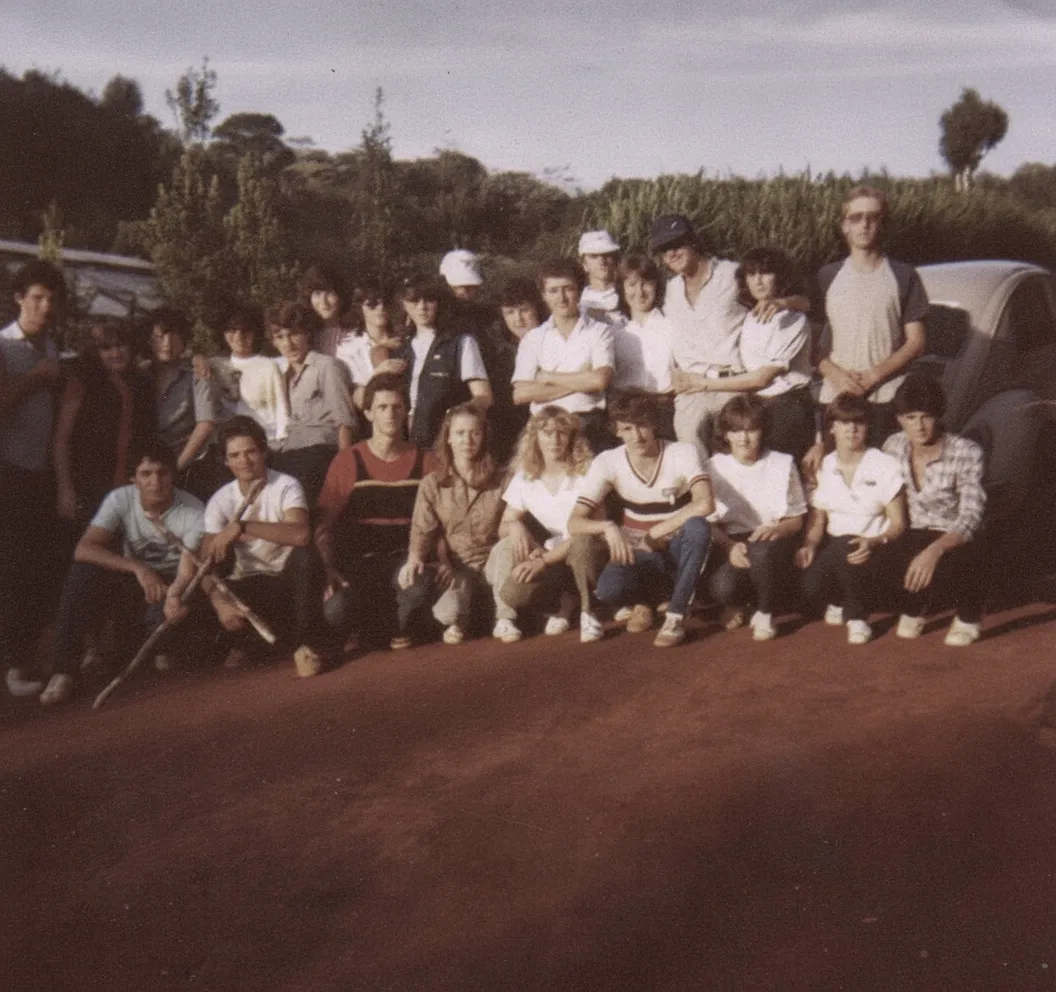 Um grupo de pessoas posando para uma foto ao ar livre, algumas sentadas e outras em pé, com árvores e um carro ao fundo.