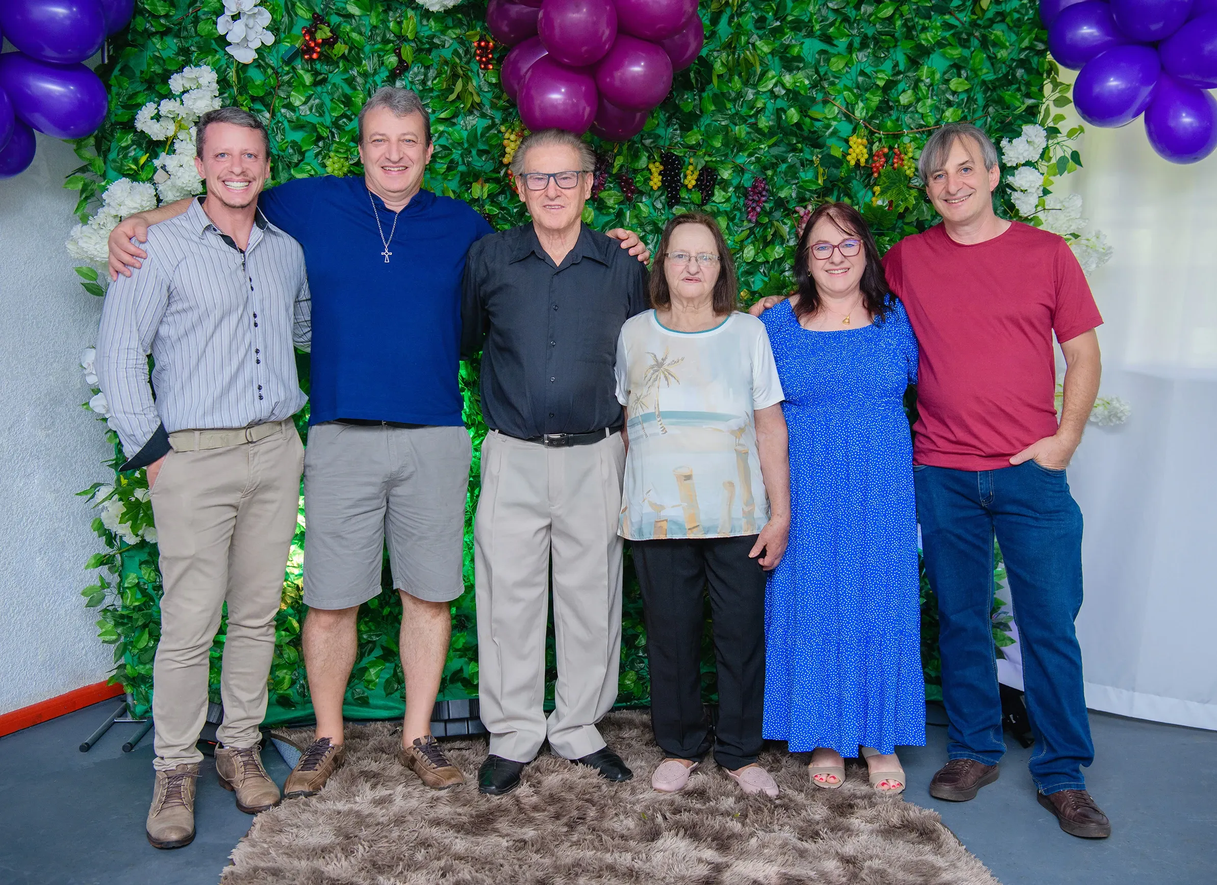 Seis adultos, três homens e três mulheres, posam para uma foto em pé, sorrindo e abraçados, em frente a uma parede de folhagem verde com algumas flores brancas e roxas, e um cacho de balões roxos no canto superior esquerdo e direito.