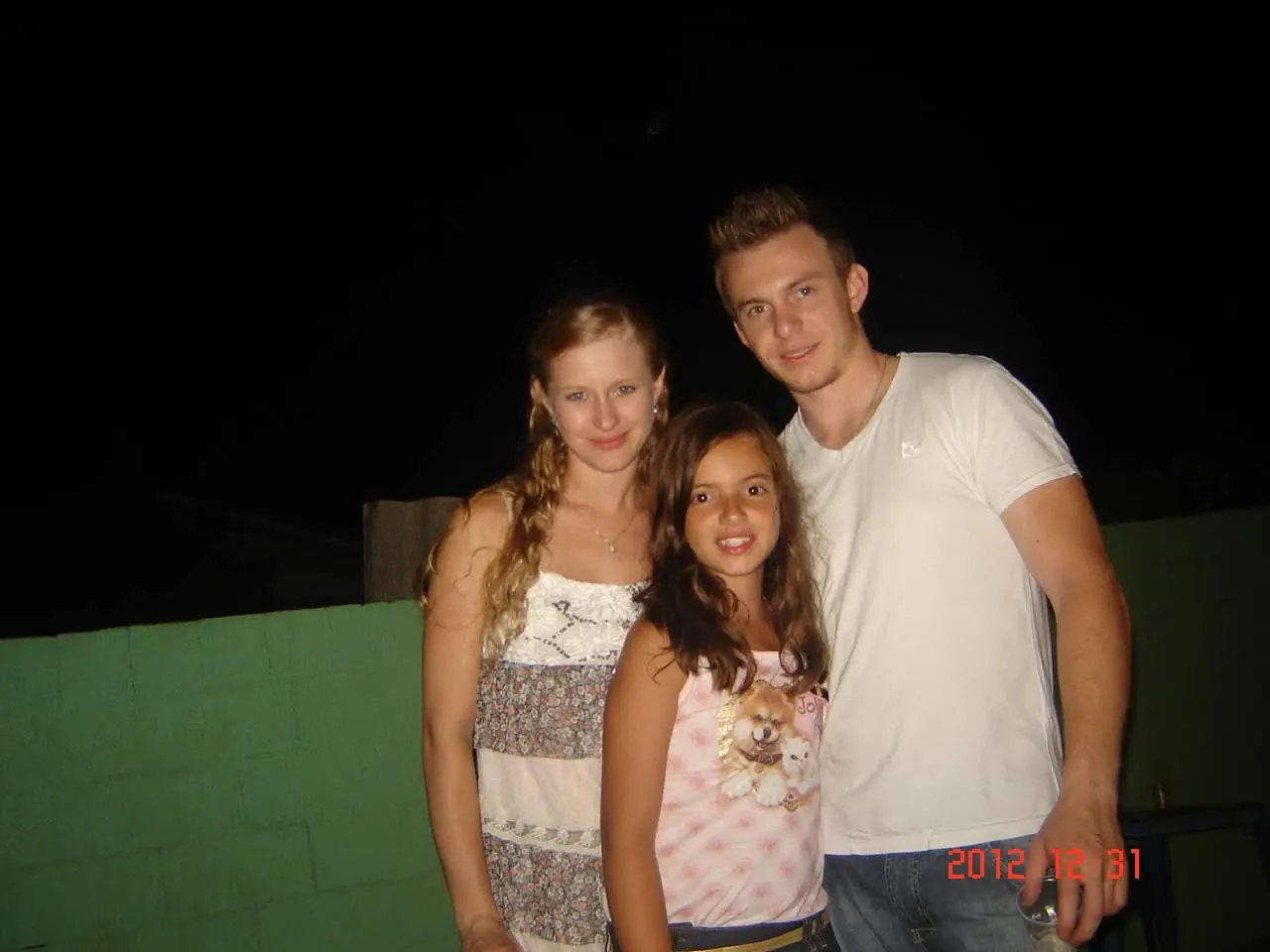 Três pessoas, duas mulheres e um homem, estão de pé e sorrindo para a câmera em frente a uma parede verde, com o fundo escuro.