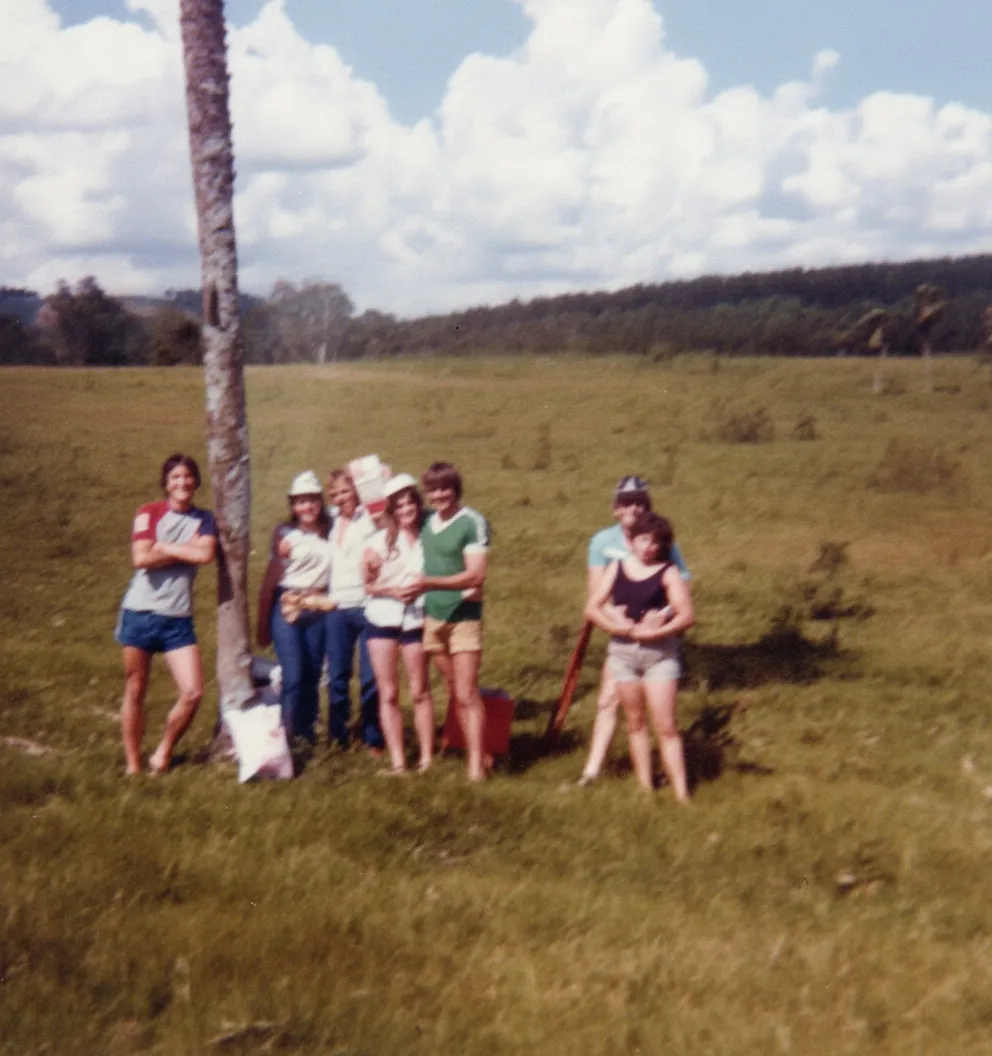 Sete jovens em roupas esportivas ficam em um campo gramado ao lado de uma única palmeira sob um céu parcialmente nublado, com colinas e árvores ao fundo.