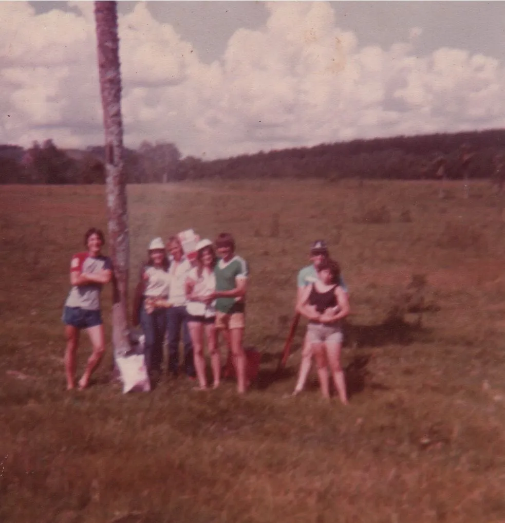Sete jovens em roupas esportivas ficam em um campo gramado ao lado de uma única palmeira sob um céu parcialmente nublado, com colinas e árvores ao fundo. (Original)