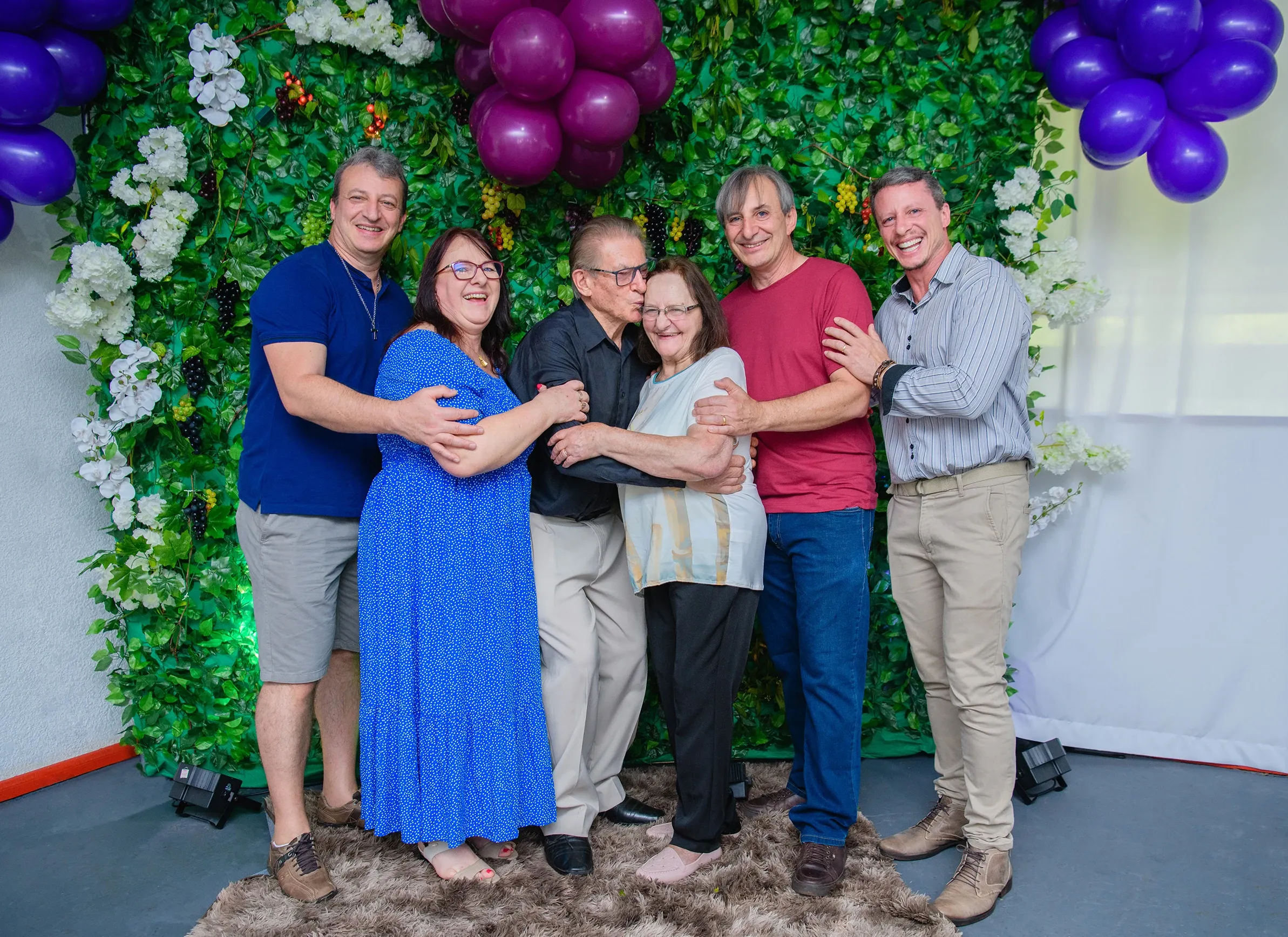 Seis adultos, três homens e três mulheres, posam para uma foto em frente a um pano de fundo de folhas e flores verdes, com balões roxos no topo.