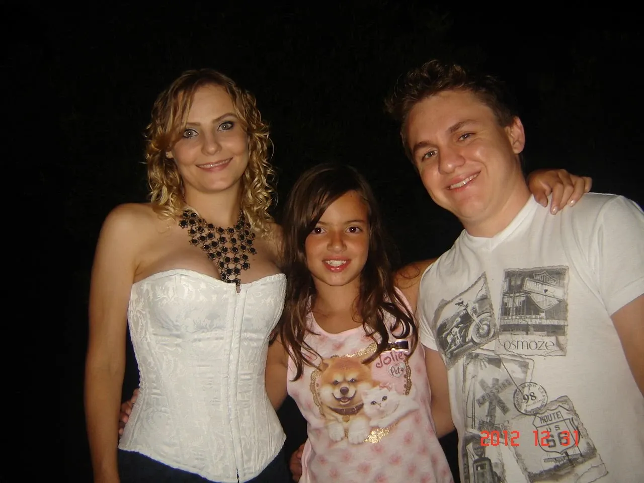 Três pessoas, uma mulher de cabelo loiro e espartilho branco com colar preto, uma menina de cabelo castanho e camisa rosa estampada, e um homem de cabelo loiro e camiseta branca estampada, posam lado a lado e sorriem para a câmera em um ambiente escuro, com um carimbo de data "2012 6 6" no canto inferior direito.