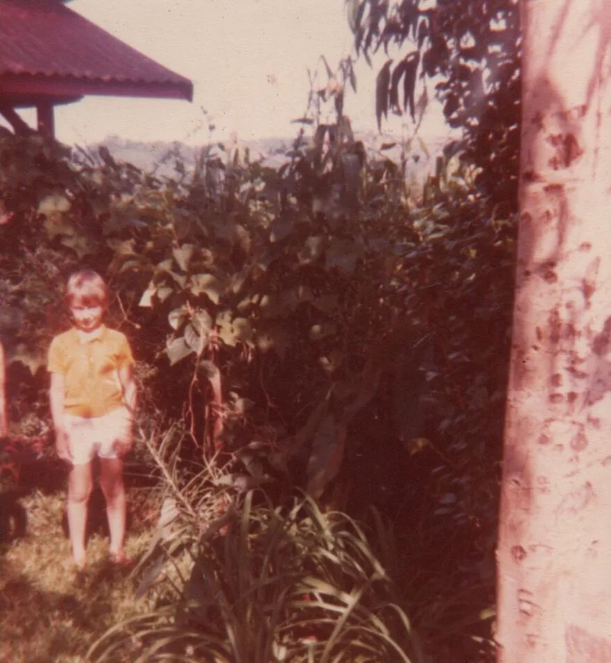 Menino loiro com camisa polo amarela e shorts brancos, sorrindo e olhando para a câmera, de pé em um jardim verdejante com um pequeno telhado vermelho e uma árvore no fundo. (Original)