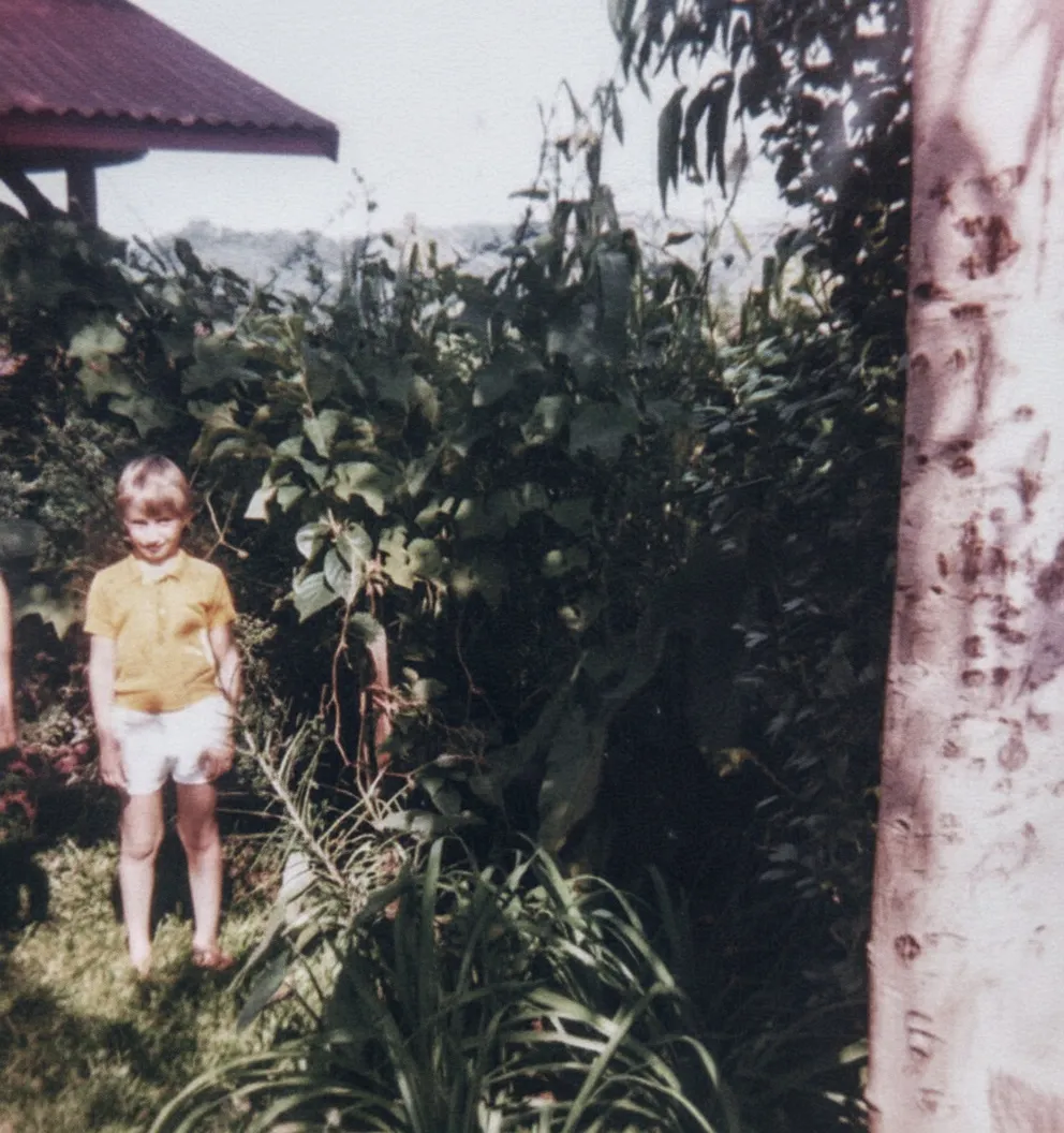 Menino loiro com camisa polo amarela e shorts brancos, sorrindo e olhando para a câmera, de pé em um jardim verdejante com um pequeno telhado vermelho e uma árvore no fundo.