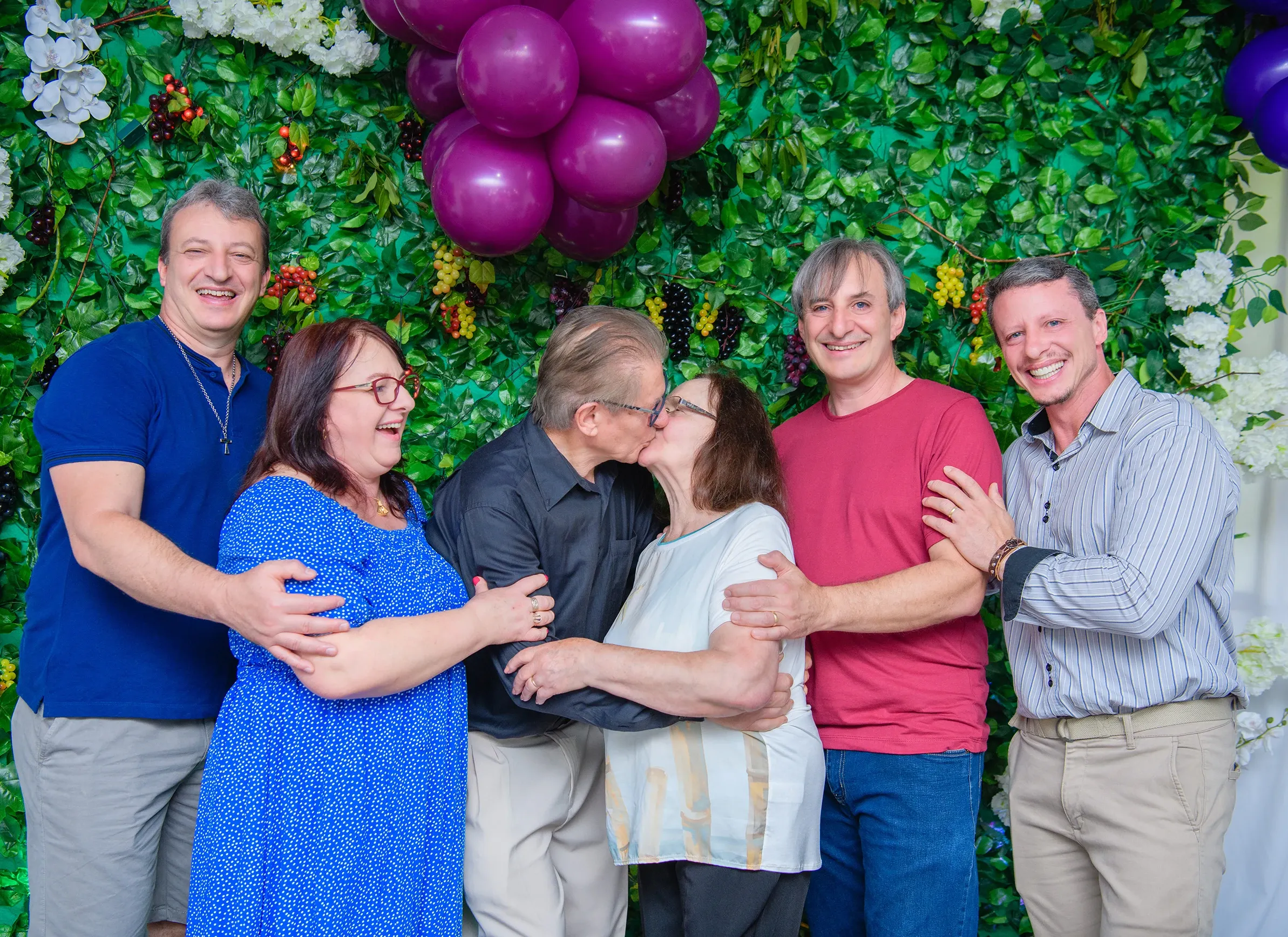 Seis adultos de meia-idade e mais velhos posam para uma foto em frente a uma parede coberta por folhagem verde e cachos de uvas, com balões roxos no canto superior direito; os dois no centro se beijam, enquanto os outros quatro os abraçam e sorriem para a câmera.