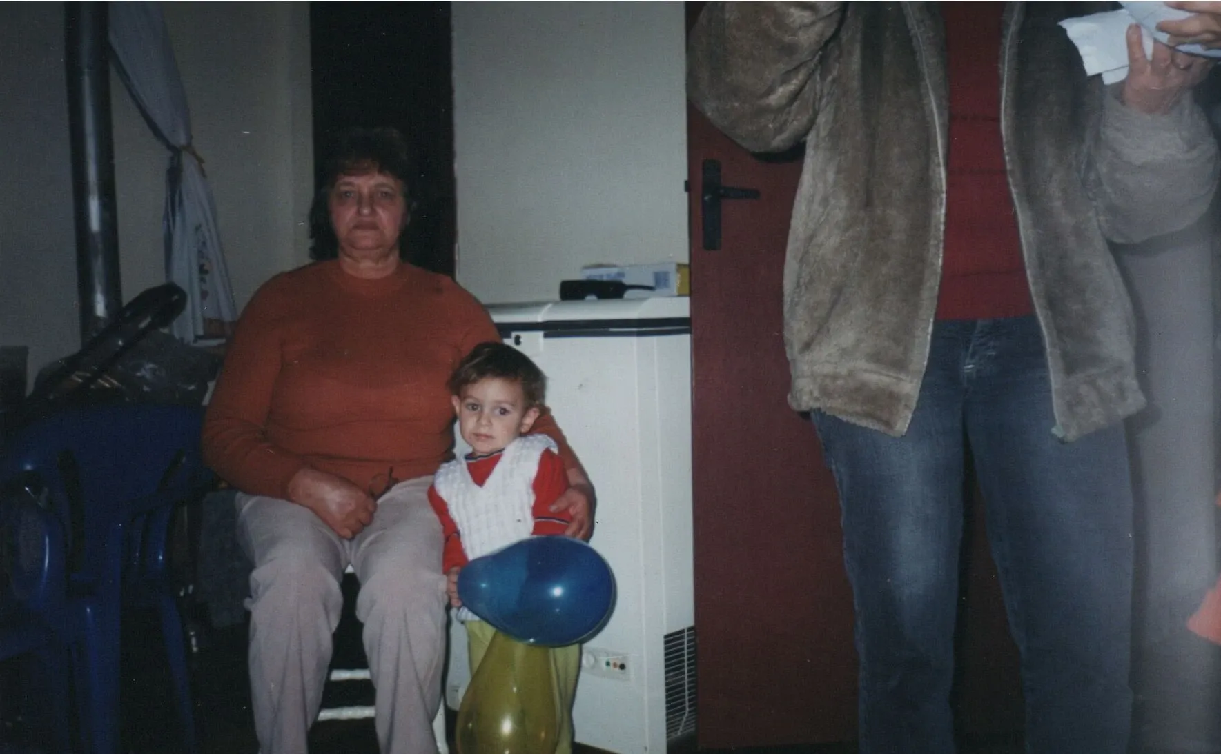 Uma avó sentada, vestindo um suéter laranja, segura um menino pequeno que está em pé à sua frente, segurando um balão azul e amarelo; uma mulher cortada está à direita da imagem.