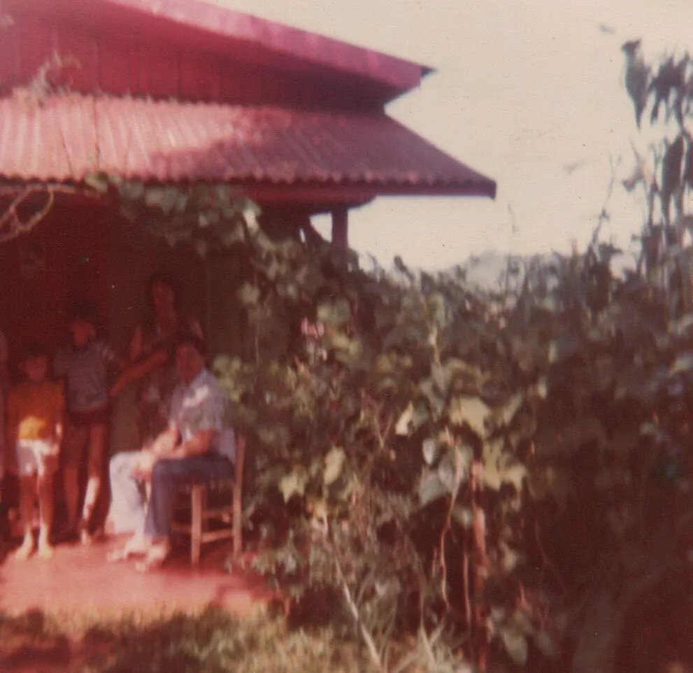 Pessoas estão sentadas e em pé na varanda de uma casa de um andar, com um telhado de telha vermelha. (Original)