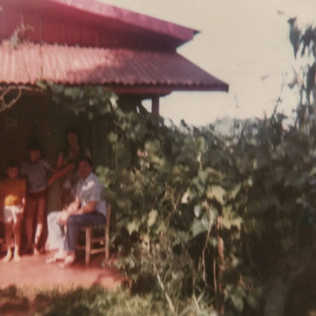 Pessoas estão sentadas e em pé na varanda de uma casa de um andar, com um telhado de telha vermelha.