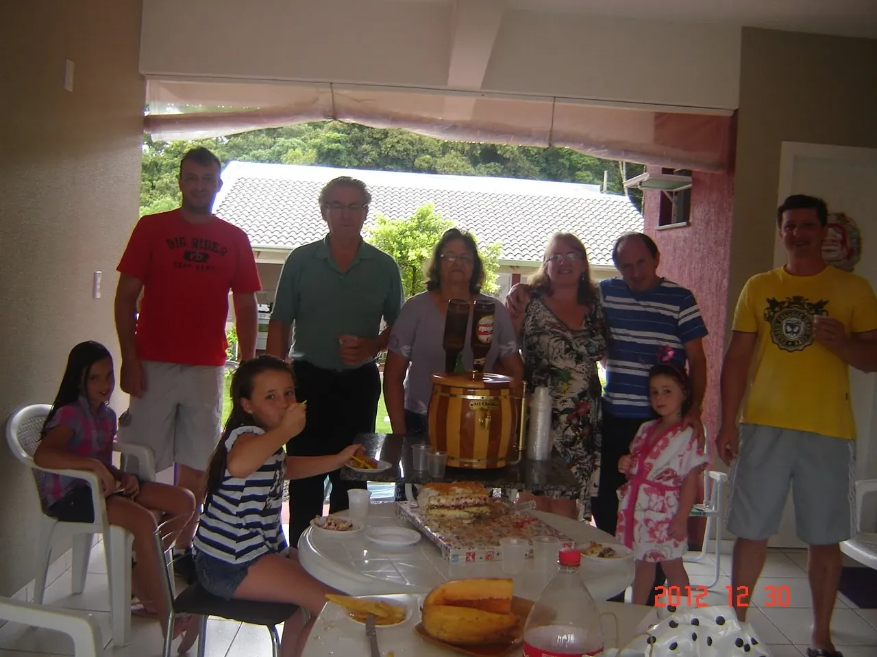 Um grupo de oito pessoas, incluindo duas crianças, posa para uma foto em um pátio com uma mesa de comida e bebidas.