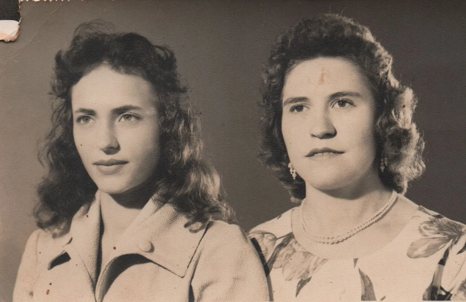 Duas mulheres, uma com cabelo castanho encaracolado e um casaco bege, e outra com cabelo encaracolado mais escuro, um colar de pérolas e um vestido floral, posam para um retrato. (Original)