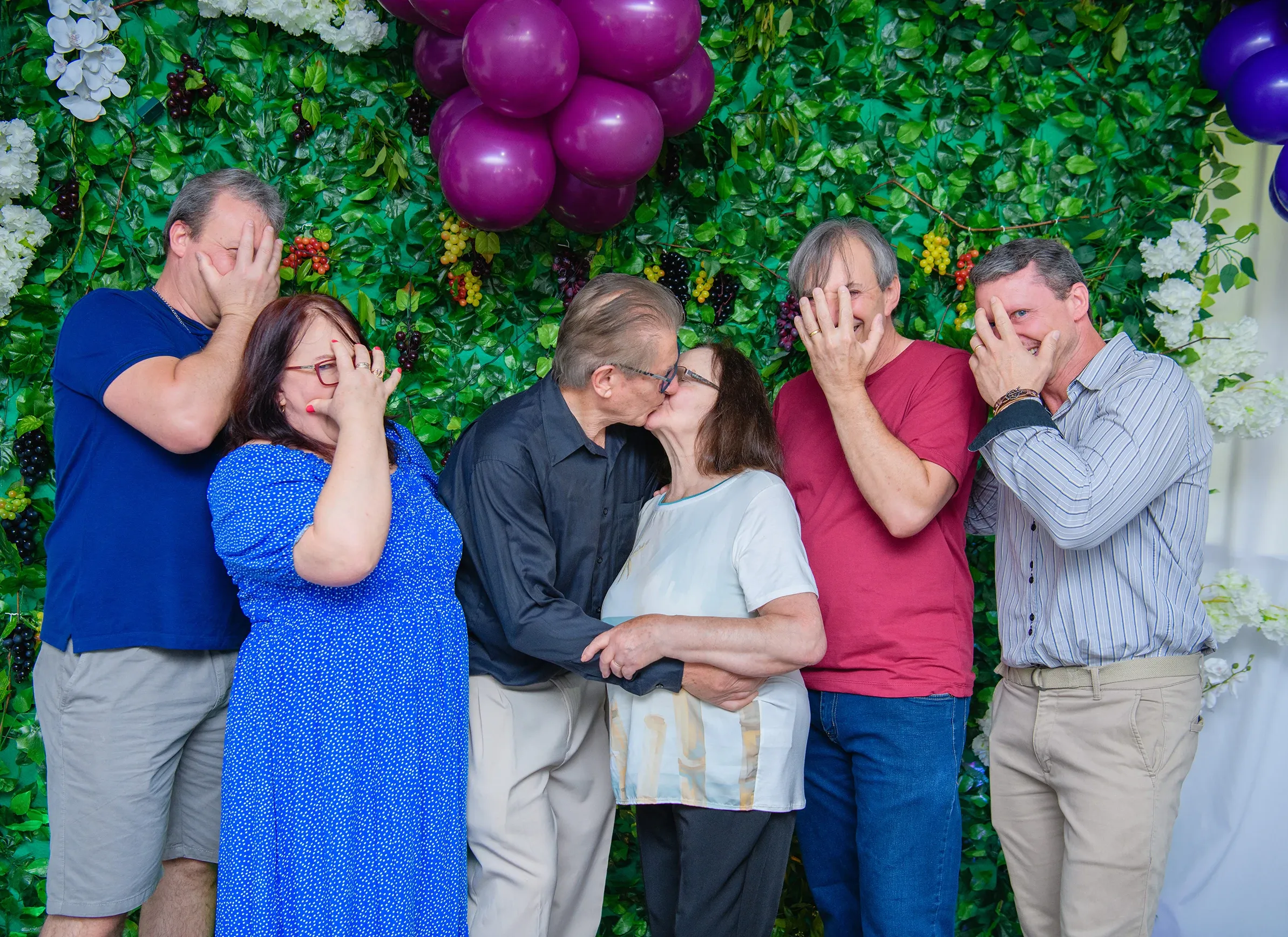 Um casal mais velho se beija no centro, com três homens e uma mulher de cada lado cobrindo os rostos com as mãos, em frente a uma parede de vegetação e balões roxos.