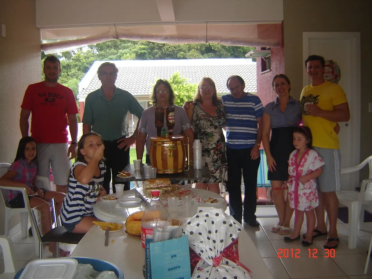 Um grupo de dez pessoas, incluindo adultos e crianças, posa para uma foto em um encontro familiar em uma varanda com uma mesa de comida e bebidas.