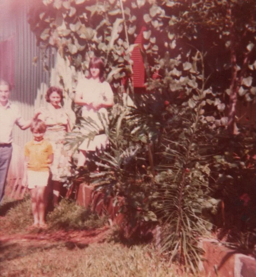 Quatro pessoas em um jardim, com um homem de pé no canto esquerdo, duas mulheres no meio, e uma criança no canto inferior esquerdo, todos cercados por folhagem. (Original)