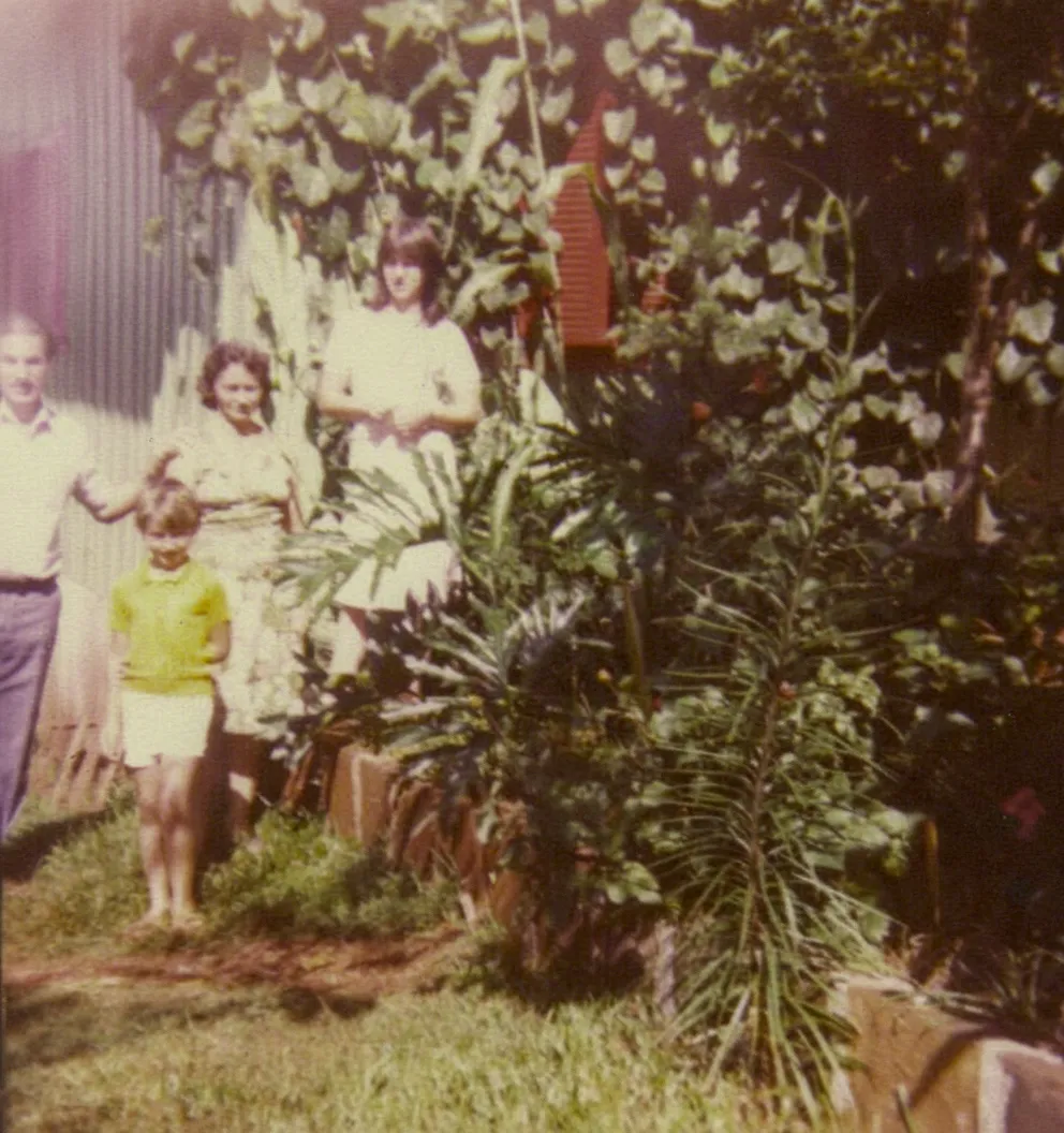 Quatro pessoas em um jardim, com um homem de pé no canto esquerdo, duas mulheres no meio, e uma criança no canto inferior esquerdo, todos cercados por folhagem.