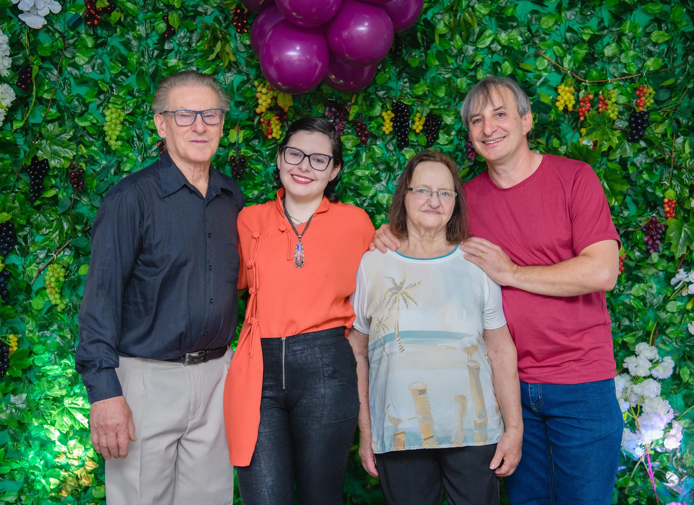 Quatro pessoas, duas mulheres e dois homens, de pé e sorrindo na frente de uma parede verde decorada com frutas e flores; um homem idoso está à esquerda, uma mulher jovem está ao lado dele, uma mulher idosa está na frente dela, e um homem de meia-idade está à direita da mulher idosa.