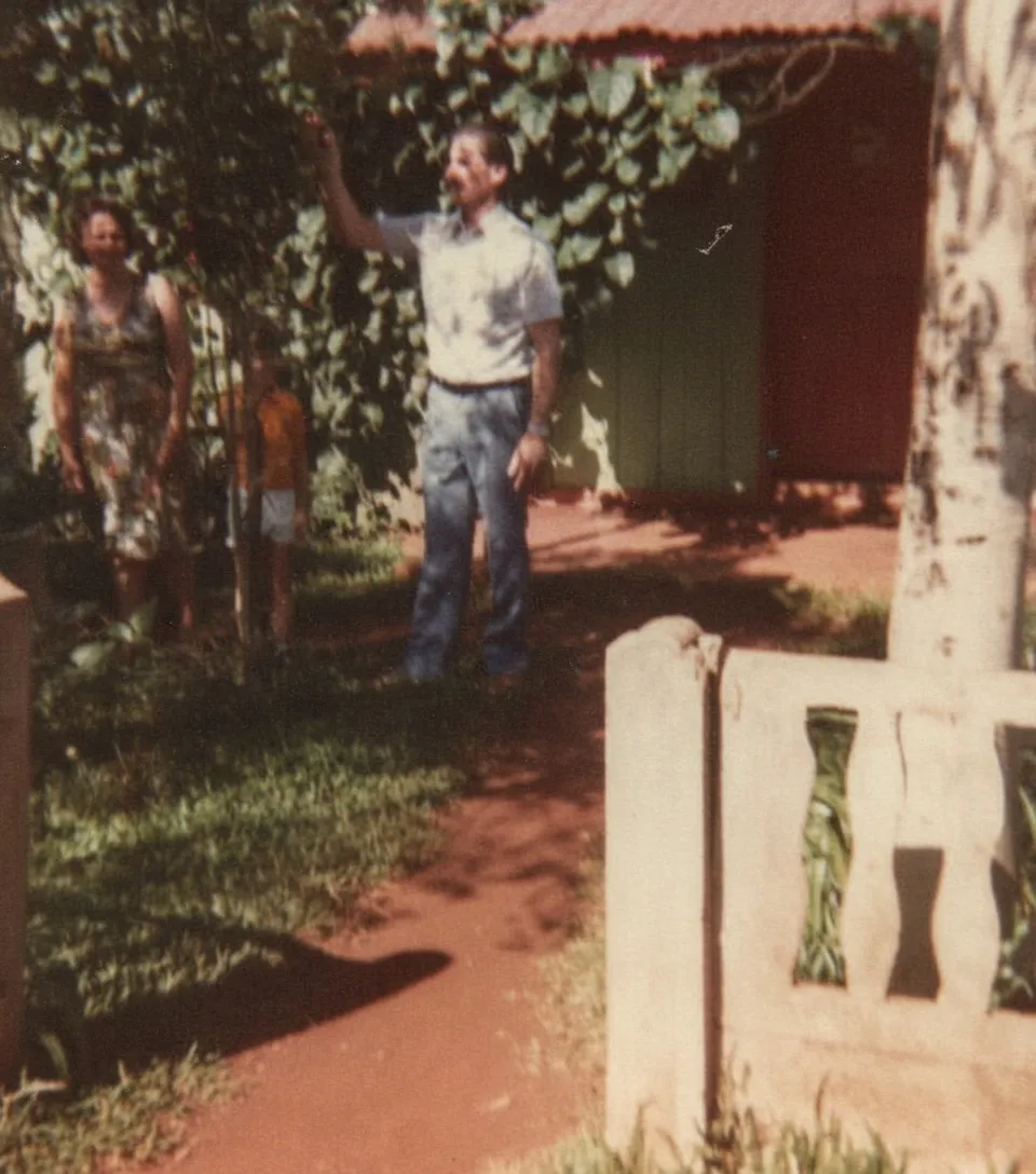 Um homem de camisa xadrez e calças escuras aponta para uma árvore em um quintal de terra, com uma mulher e uma criança em pé sob a folhagem ao fundo, com uma casa vermelha e verde à direita.