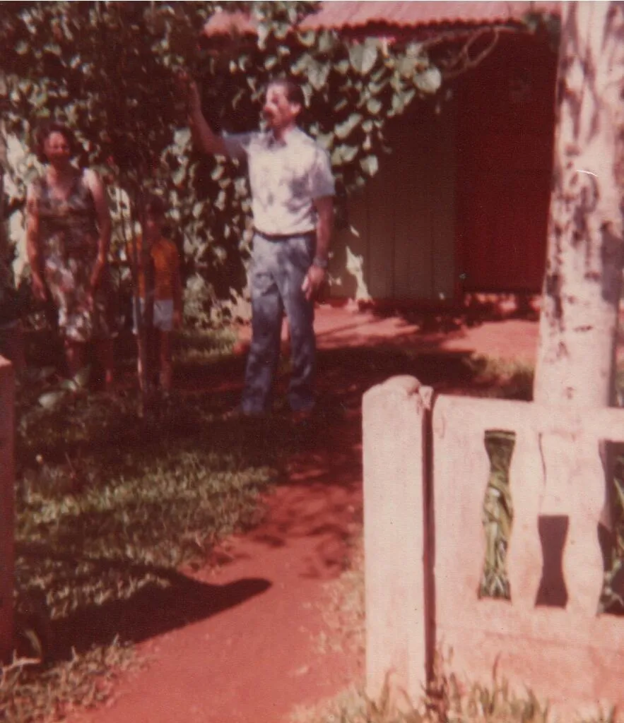 Um homem de camisa xadrez e calças escuras aponta para uma árvore em um quintal de terra, com uma mulher e uma criança em pé sob a folhagem ao fundo, com uma casa vermelha e verde à direita. (Original)