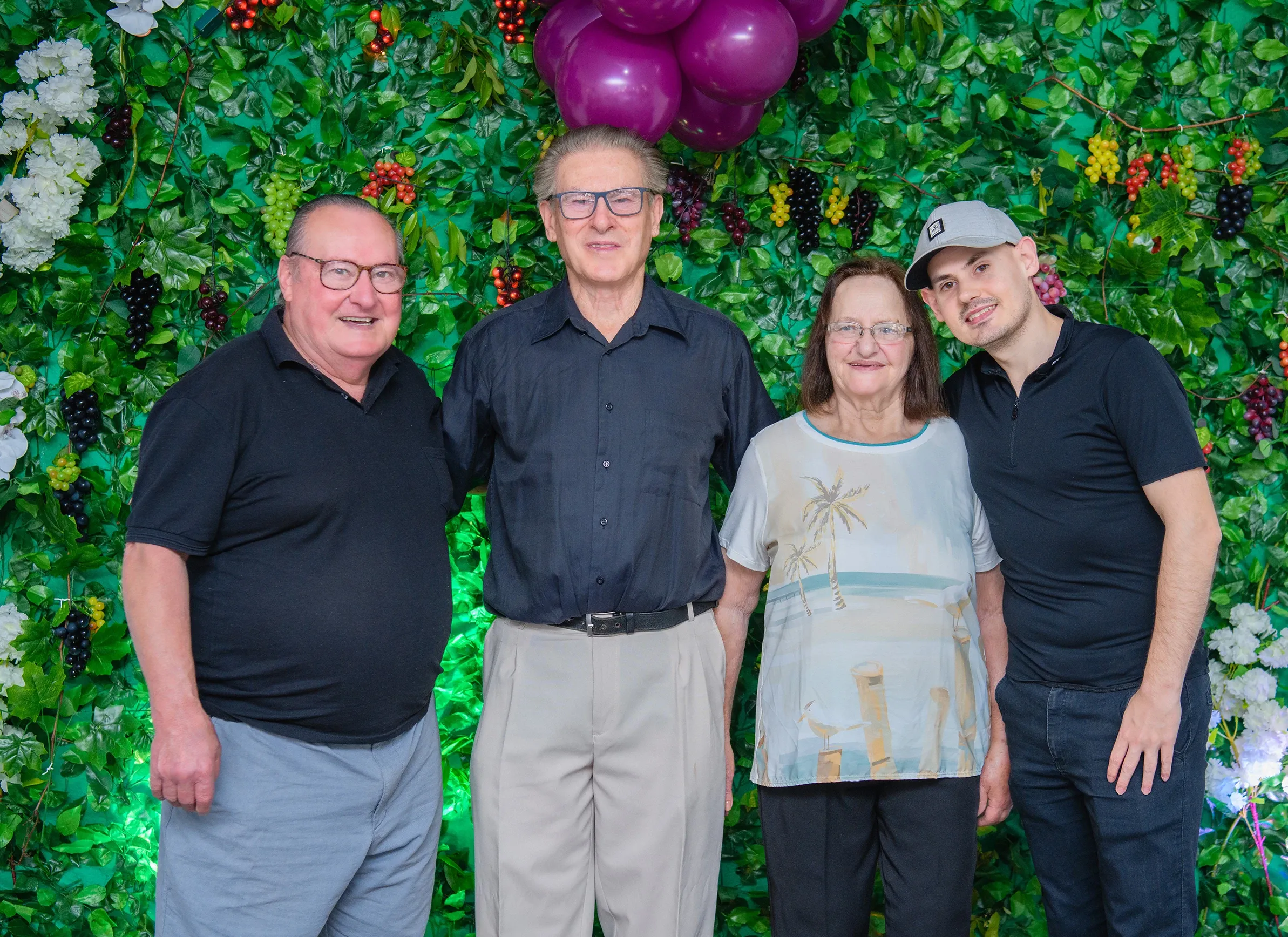 Quatro pessoas estão em pé na frente de um fundo verde exuberante feito de folhas e frutas, com dois homens idosos, uma mulher idosa e um homem mais jovem sorrindo para a câmera.