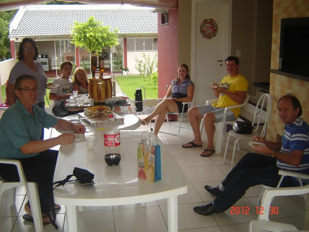 Um grupo de seis adultos e duas crianças desfrutando de uma refeição e conversando em um pátio coberto, com uma mesa de comida no centro.
