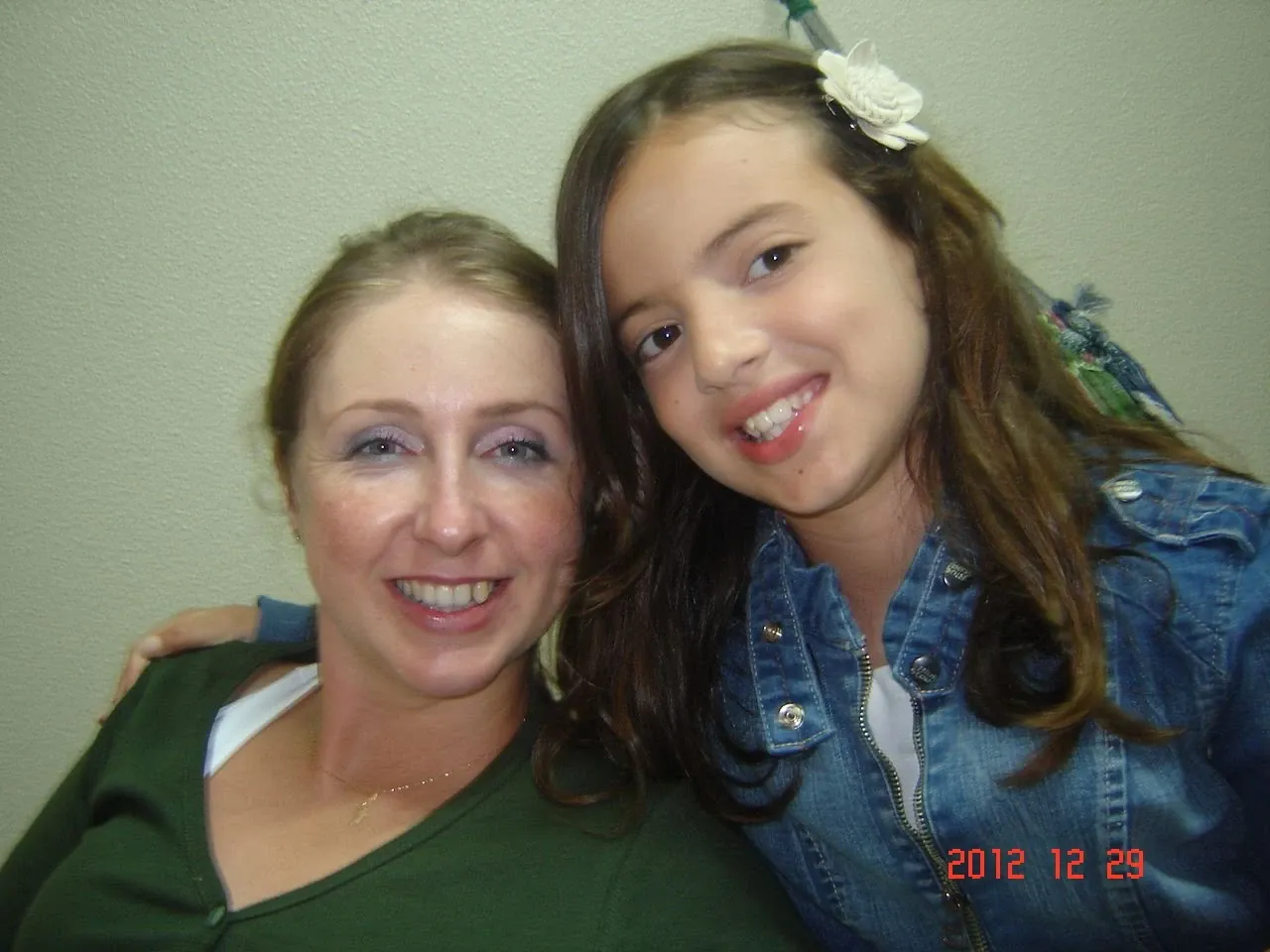 Mulher branca de meia-idade e menina sorrindo para a câmera, com a mulher à esquerda usando uma blusa verde e a menina à direita usando uma jaqueta jeans e uma flor branca no cabelo.