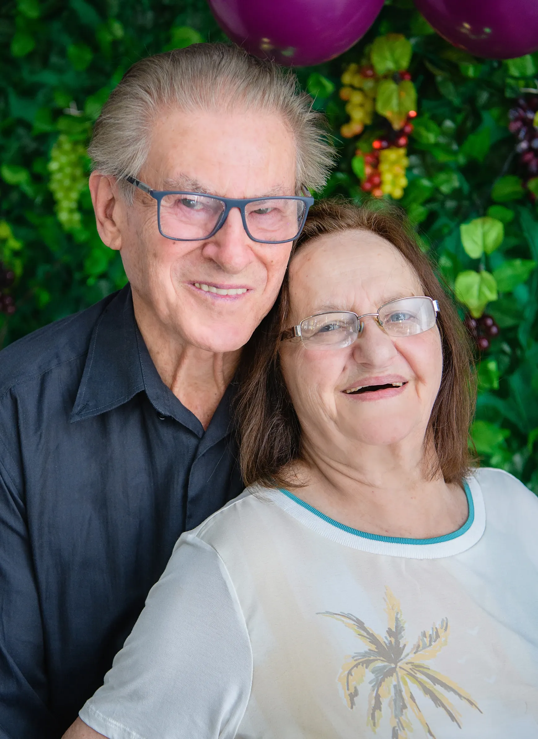 Um homem idoso com óculos e camisa escura abraçando uma mulher idosa com óculos e blusa branca, ambos sorrindo, com folhas verdes e uvas roxas e vermelhas como fundo.