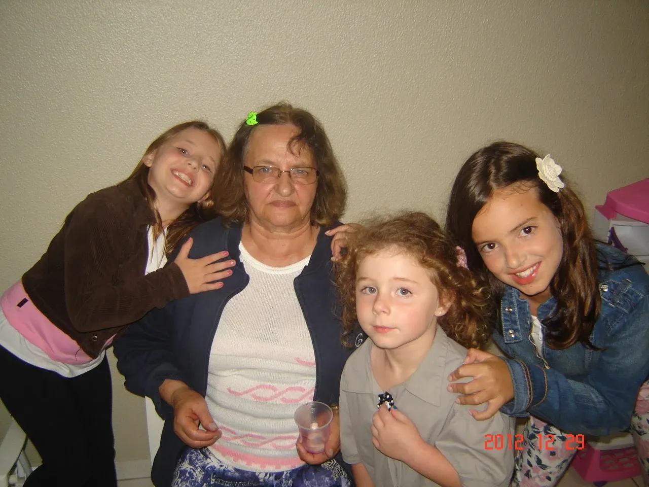 Quatro mulheres e crianças em pé e sentadas de frente para a câmera, com uma mulher mais velha no centro usando óculos e uma blusa branca e azul, ladeada por duas meninas mais novas à esquerda e à direita, e uma criança sentada no colo da mulher à frente.
