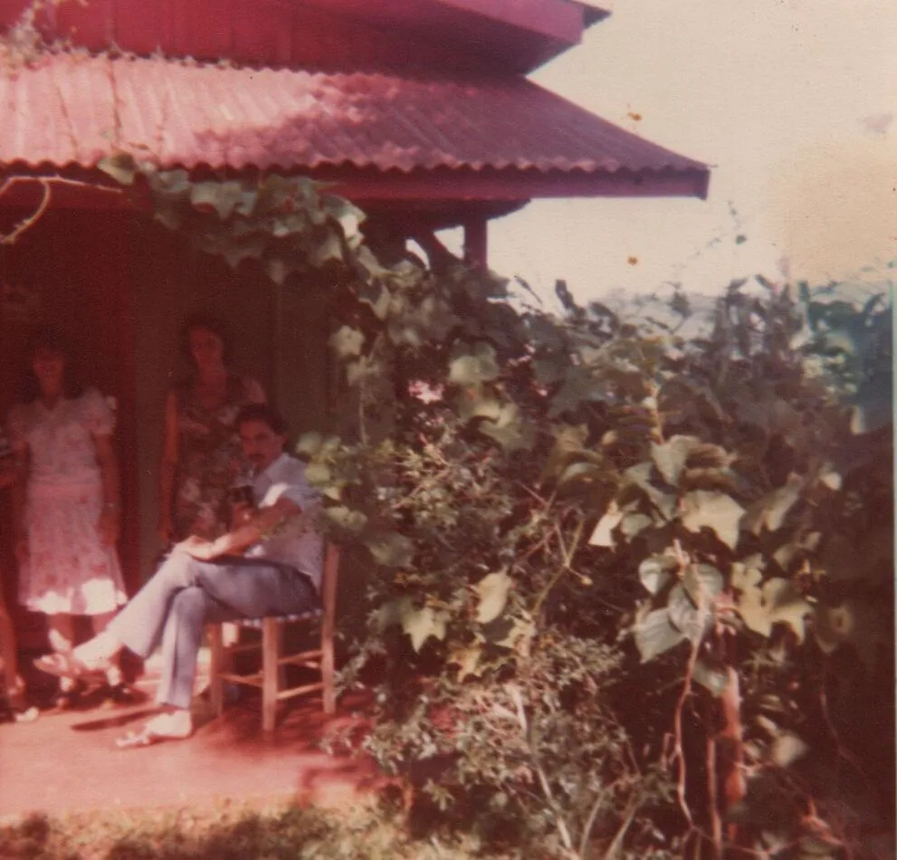 Homem sentado em cadeira de madeira do lado de fora de uma casa coberta de vinhas com três mulheres em pé ao fundo, olhando para fora da moldura. (Original)