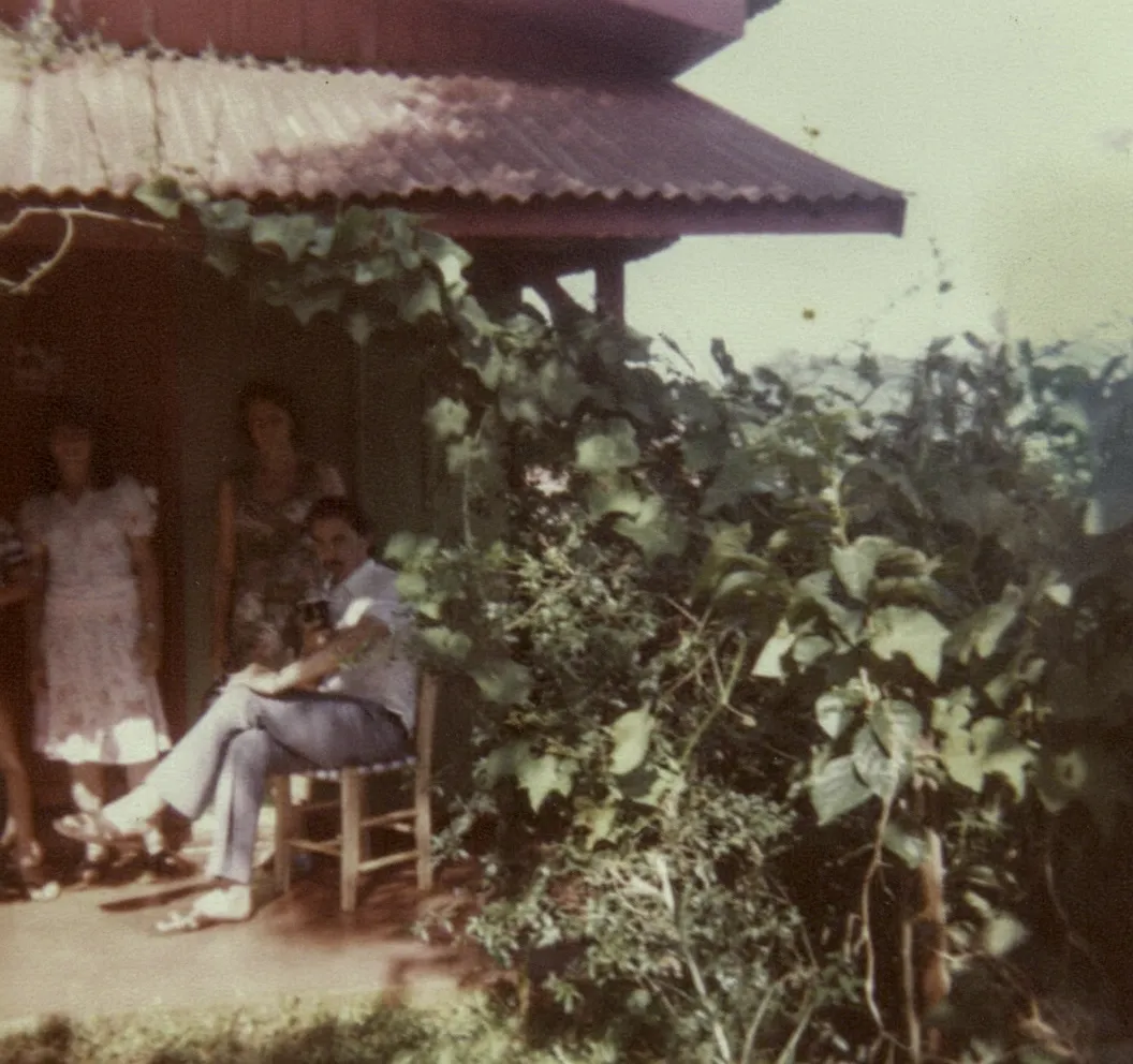 Homem sentado em cadeira de madeira do lado de fora de uma casa coberta de vinhas com três mulheres em pé ao fundo, olhando para fora da moldura.