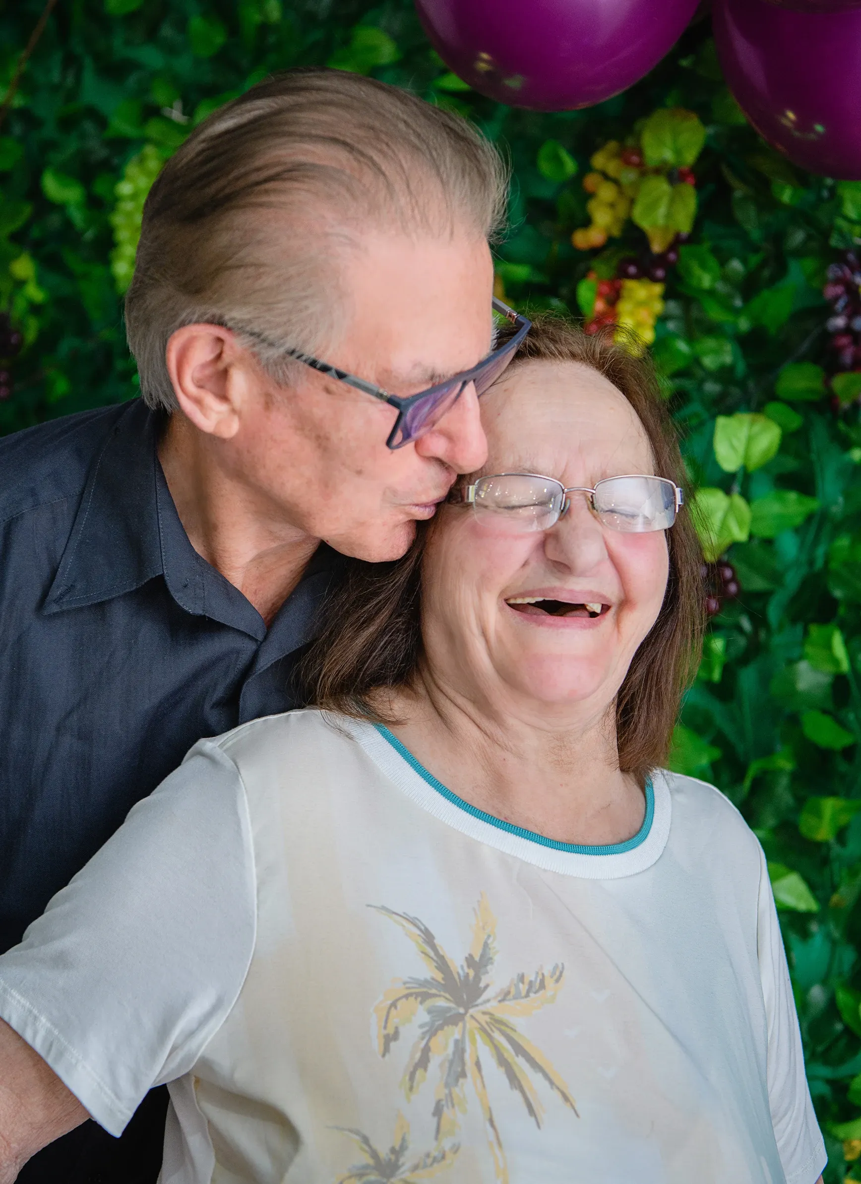 Um homem beija uma mulher no rosto, que ri em resposta, com um fundo de folhas verdes e uvas roxas e um balão roxo visível na parte superior.