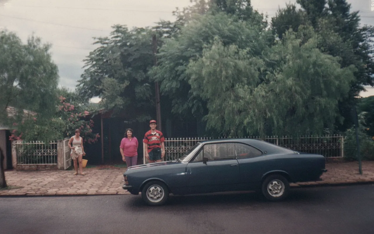 Um carro cupê azul escuro estacionado em uma rua residencial. Duas mulheres e um homem estão parados no meio-fio atrás do carro, com uma cerca de arame e árvores exuberantes no fundo.