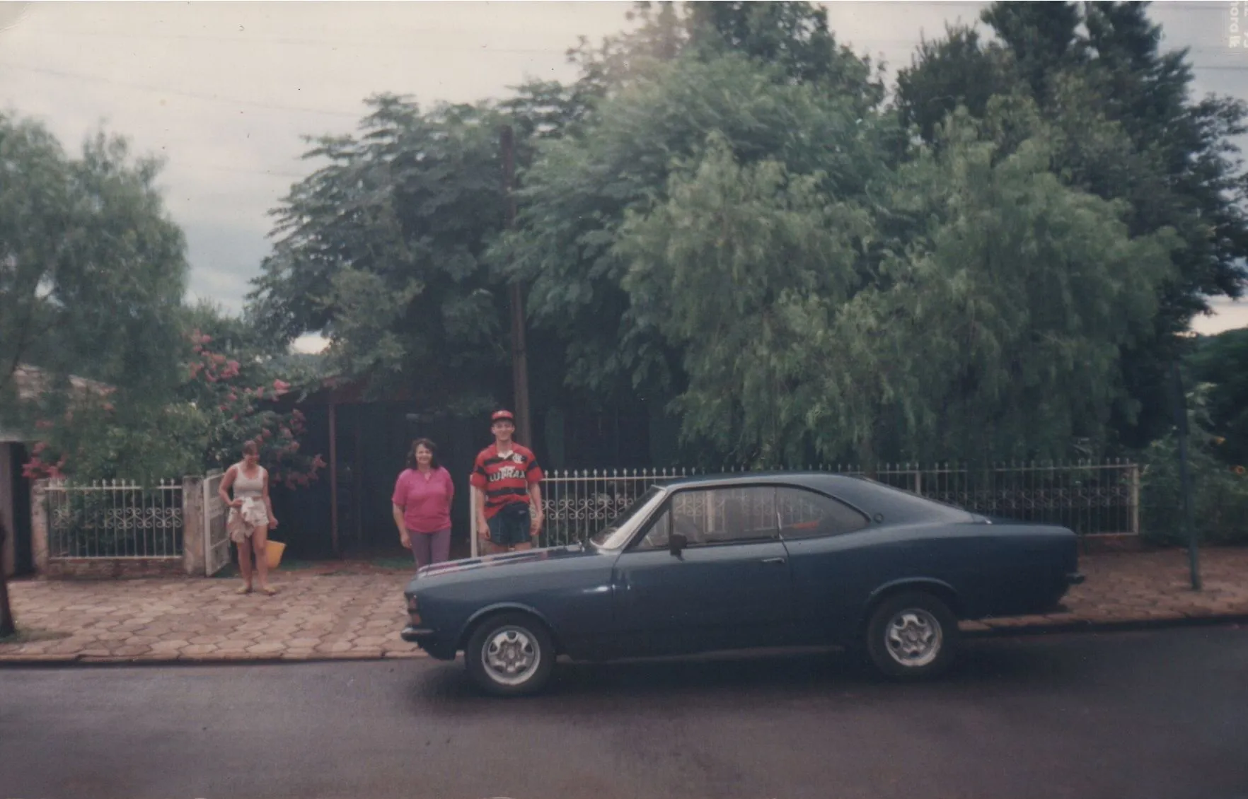Um carro cupê azul escuro estacionado em uma rua residencial. Duas mulheres e um homem estão parados no meio-fio atrás do carro, com uma cerca de arame e árvores exuberantes no fundo. (Original)