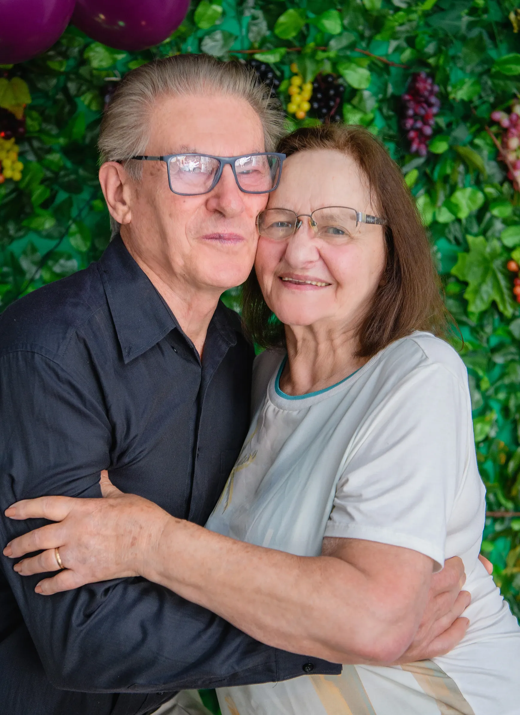 Um homem idoso com óculos e camisa azul-marinho abraça uma mulher idosa de óculos e camiseta branca, ambos sorrindo para a câmera em frente a uma parede decorada com folhagens verdes e frutas.