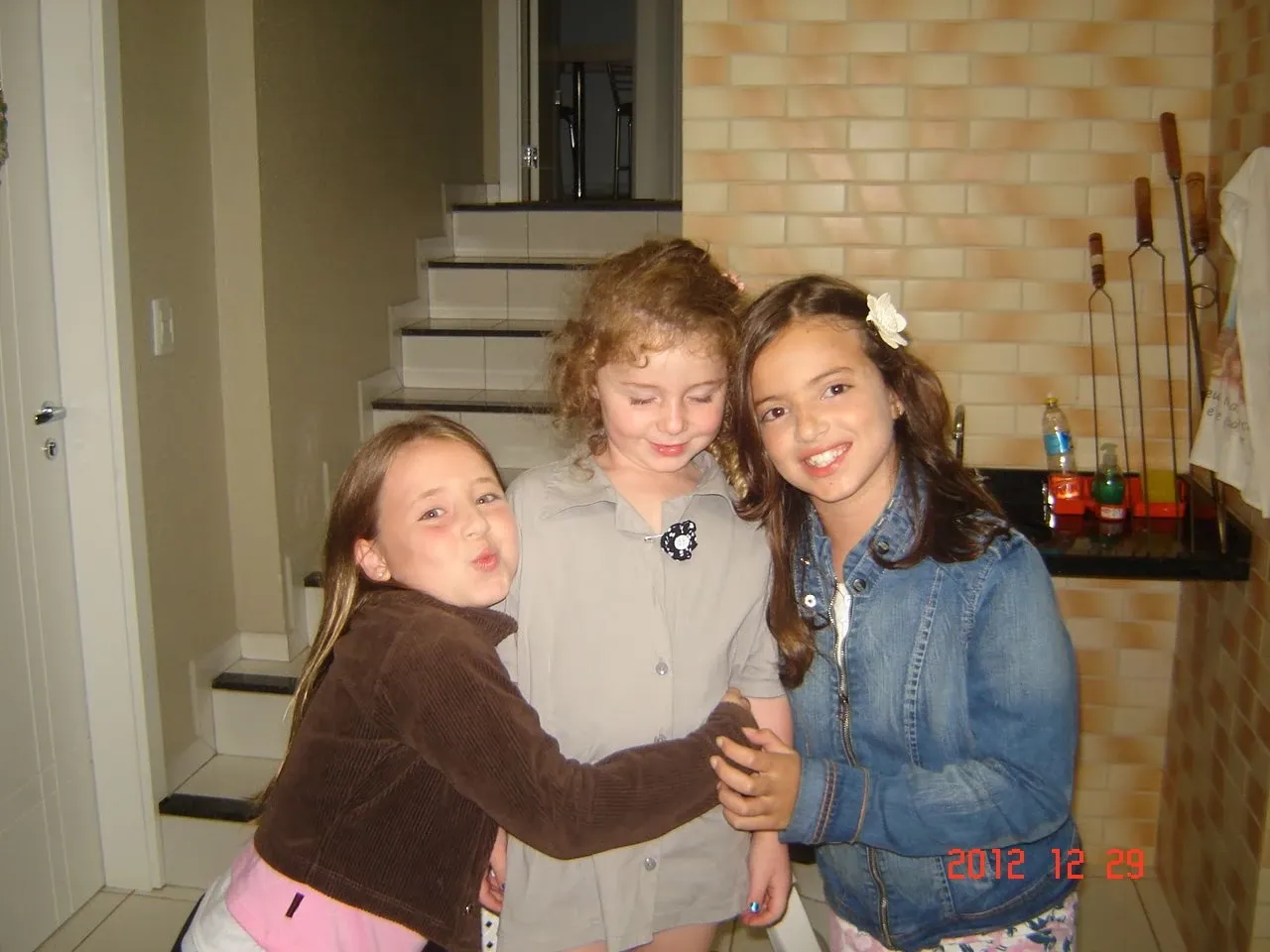 Três jovens, duas com cabelo castanho e uma com cabelo ruivo, abraçando-se e sorrindo dentro de casa, com uma delas fazendo bico para a câmera e outra usando uma flor no cabelo.