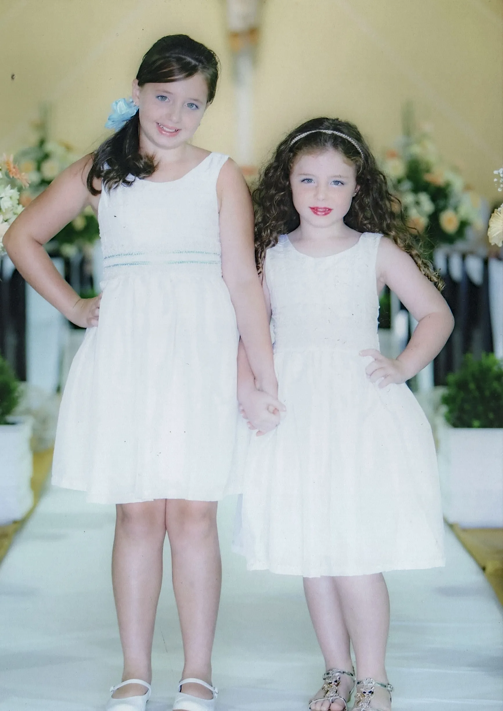 Duas meninas em vestidos brancos, de mãos dadas, em pé em um corredor, a mais velha à esquerda sorrindo para a câmera, a mais nova à direita com cabelos cacheados.