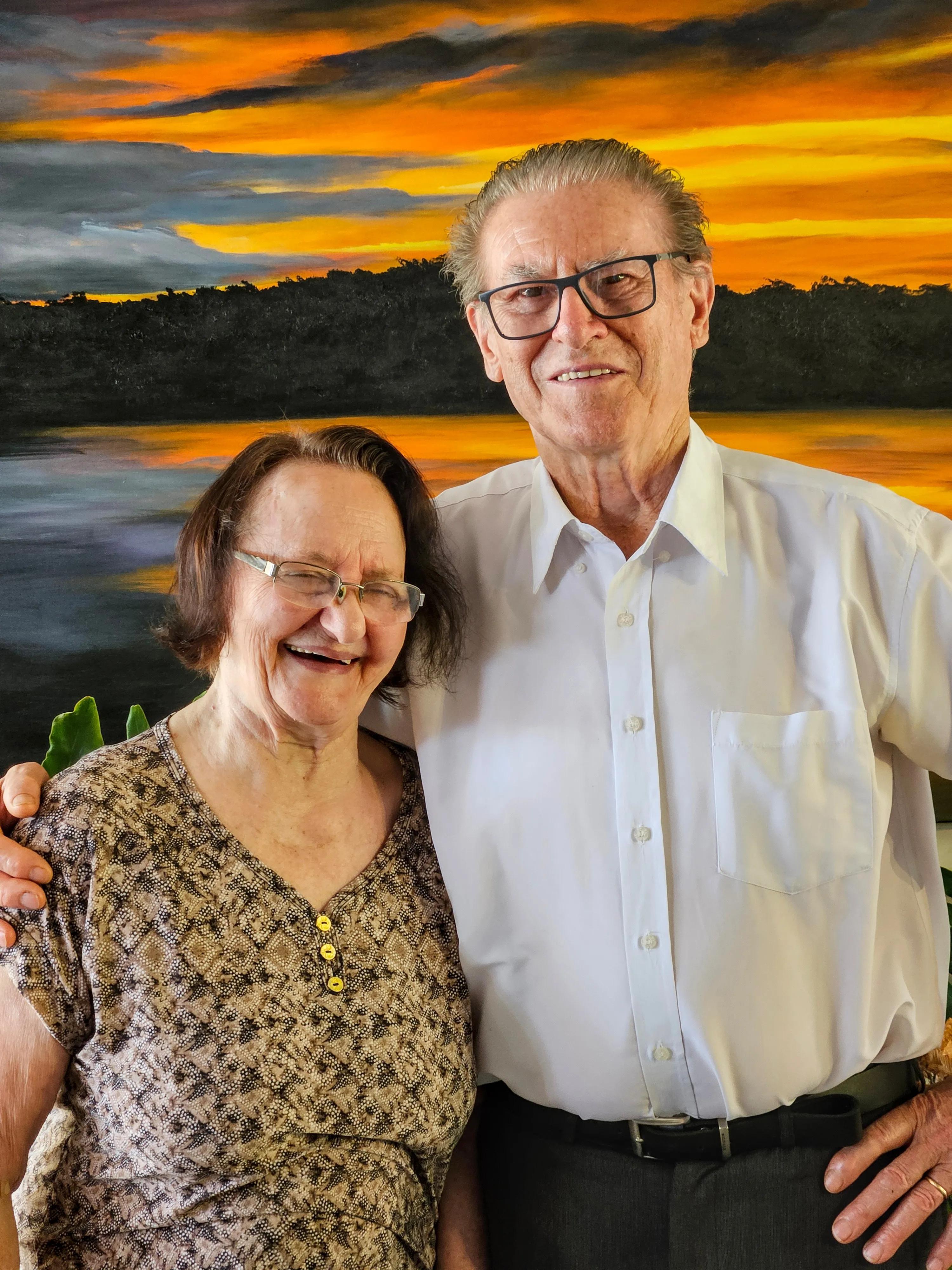 Um casal idoso sorridente, a mulher à esquerda com óculos e uma blusa estampada, o homem à direita com óculos e uma camisa social branca, abraçados na frente de uma pintura de paisagem com um pôr do sol colorido refletido em um lago.