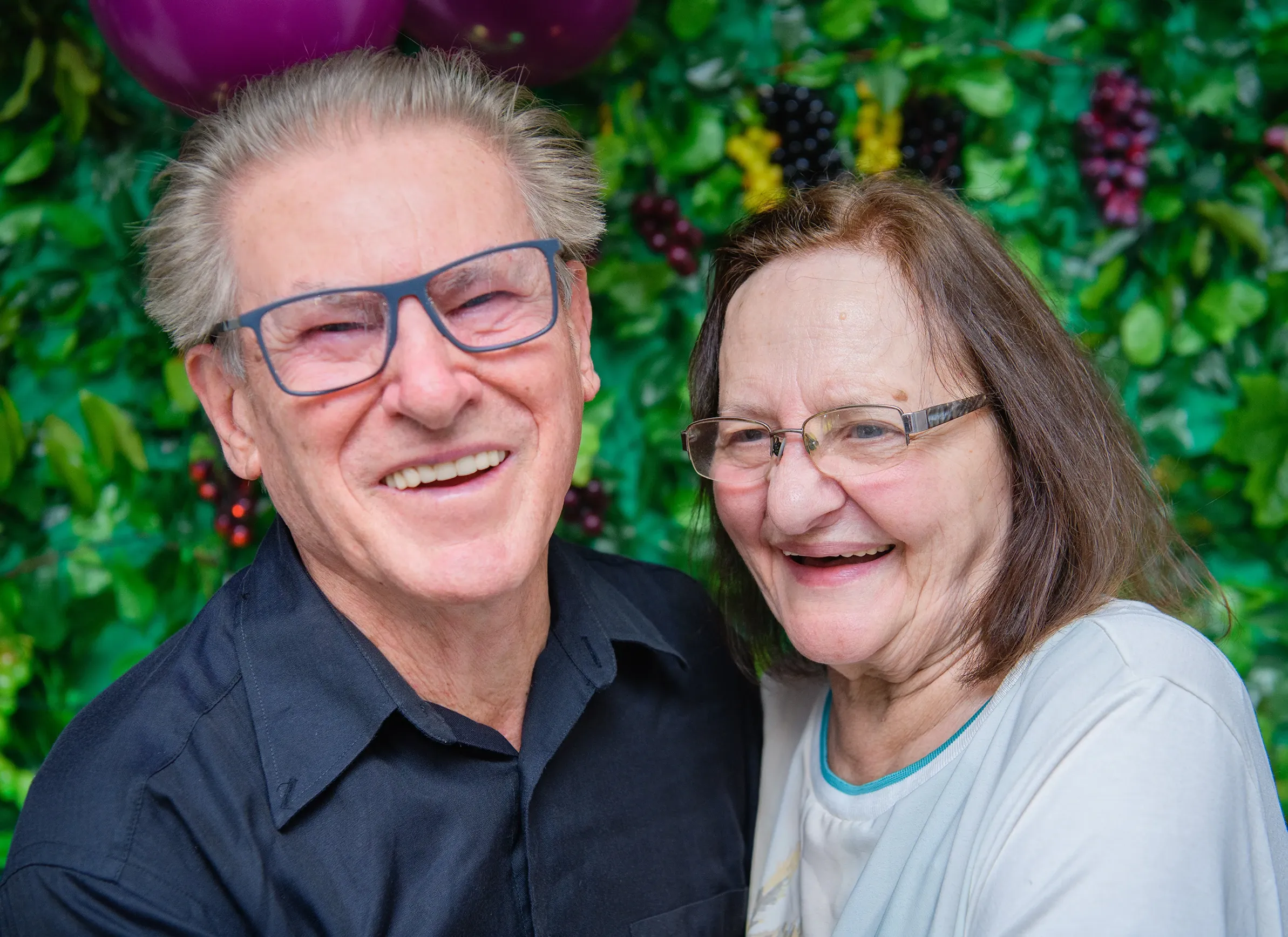 Um homem e uma mulher mais velhos, sorridentes, estão abraçados, com um fundo verde exuberante com balões roxos e frutas escuras visíveis.