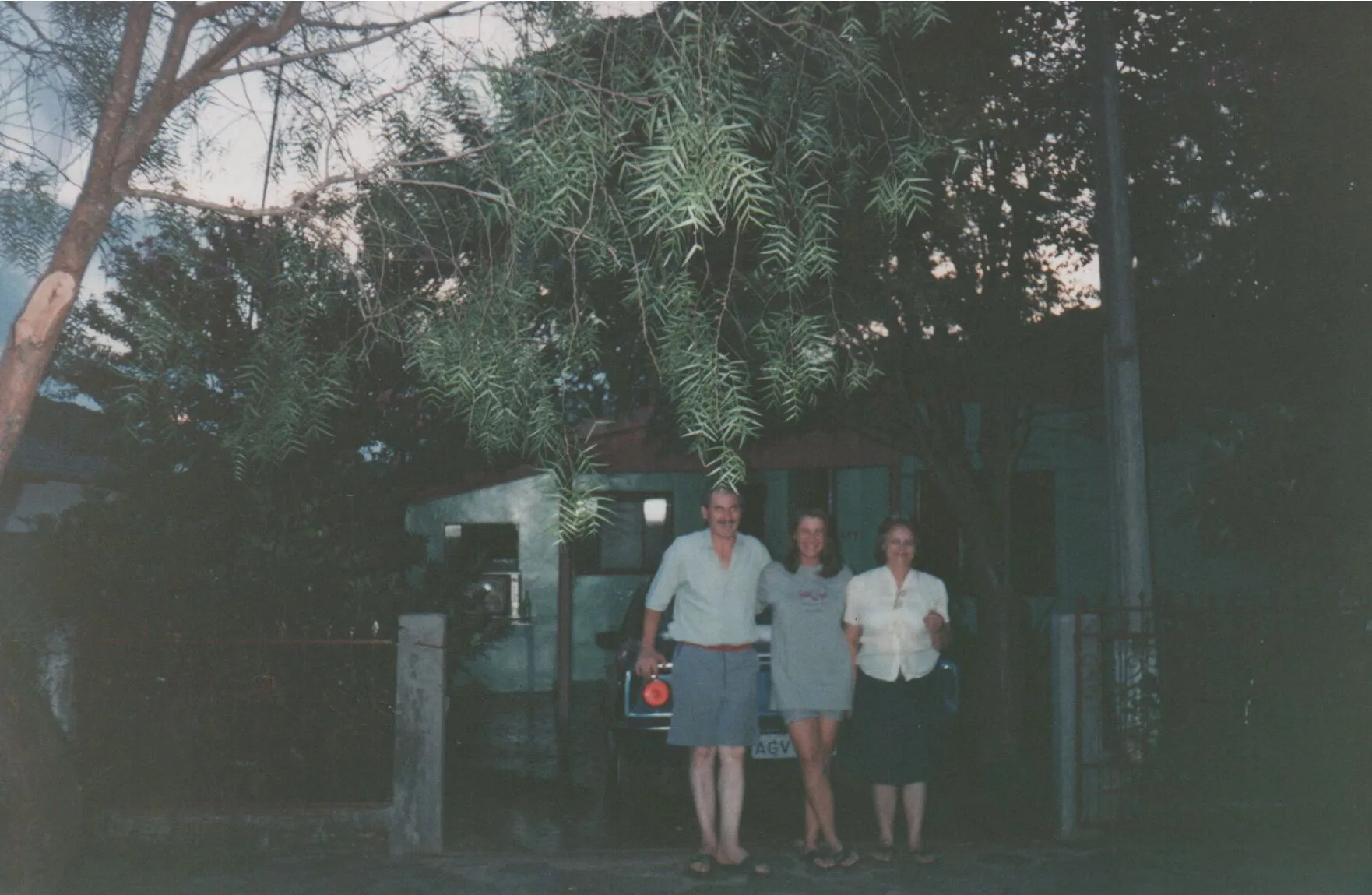 Três pessoas, duas mulheres e um homem, em pé na rua em frente a uma casa com árvores. (Original)
