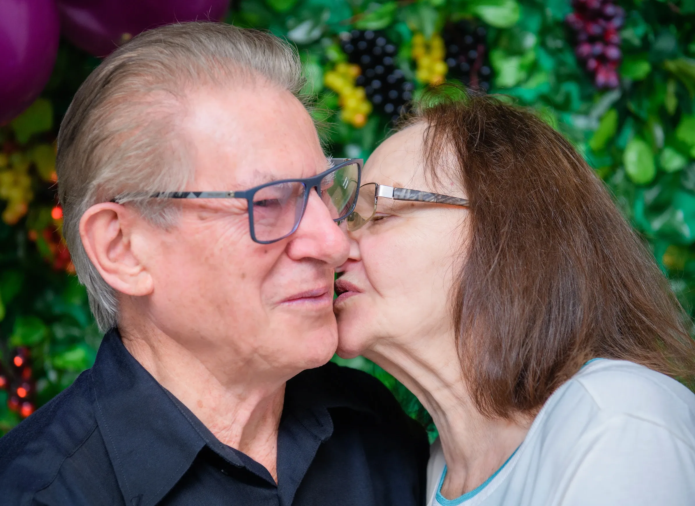 Uma mulher beija a bochecha de um homem que usa óculos, com um fundo desfocado de balões e folhagens.