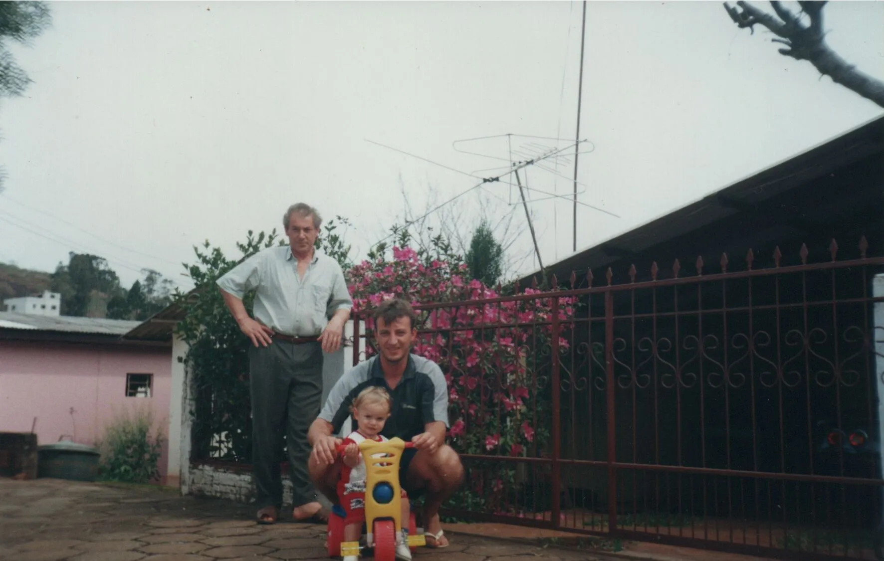 Dois homens e uma criança em um triciclo posam em frente a uma casa com uma cerca. (Original)