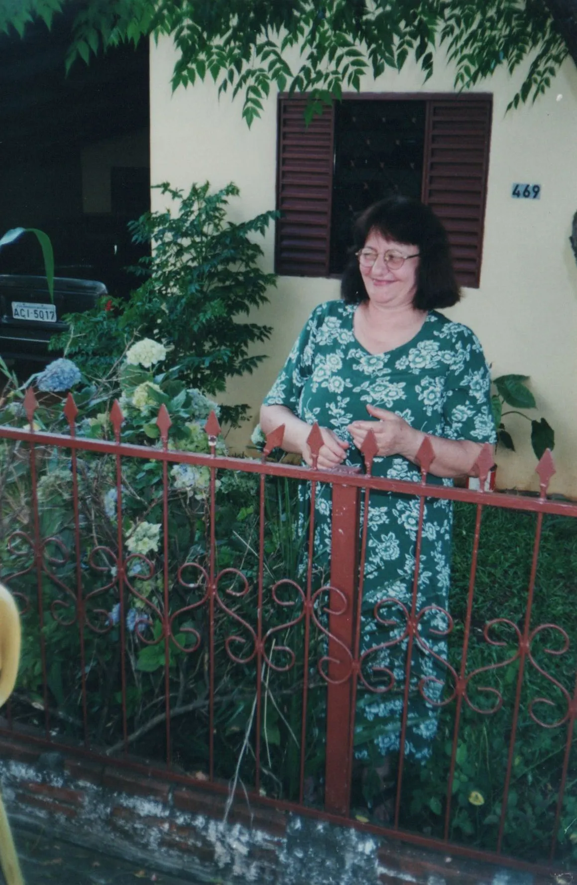 Mulher de óculos, com cabelo escuro e camisa floral verde, sorrindo atrás de uma cerca de ferro vermelha em frente a uma casa. (Original)