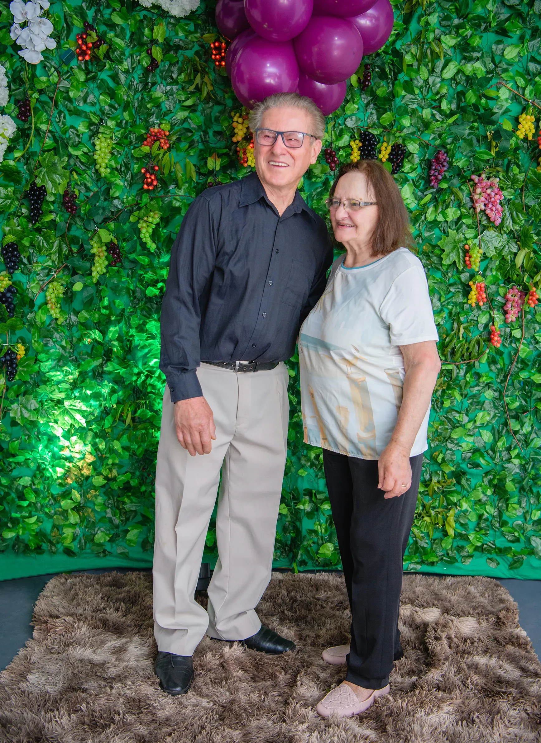 Um homem e uma mulher mais velhos, sorridentes, de pé na frente de uma parede verde decorada com folhagens e frutas, e balões roxos no topo.